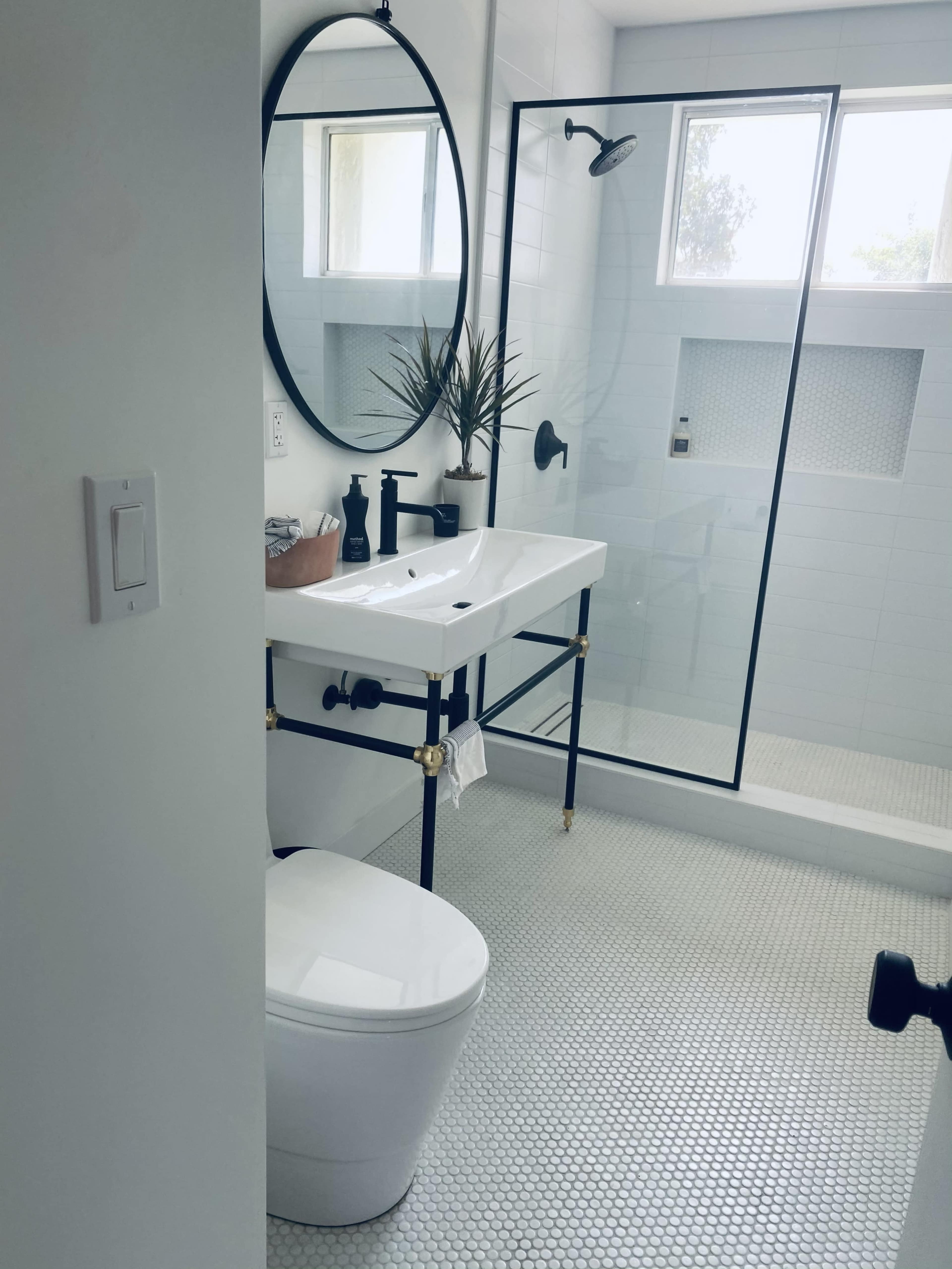 The image features a modern bathroom with a glass shower, a white pedestal sink with a circular mirror above it, a potted plant, and a toilet.