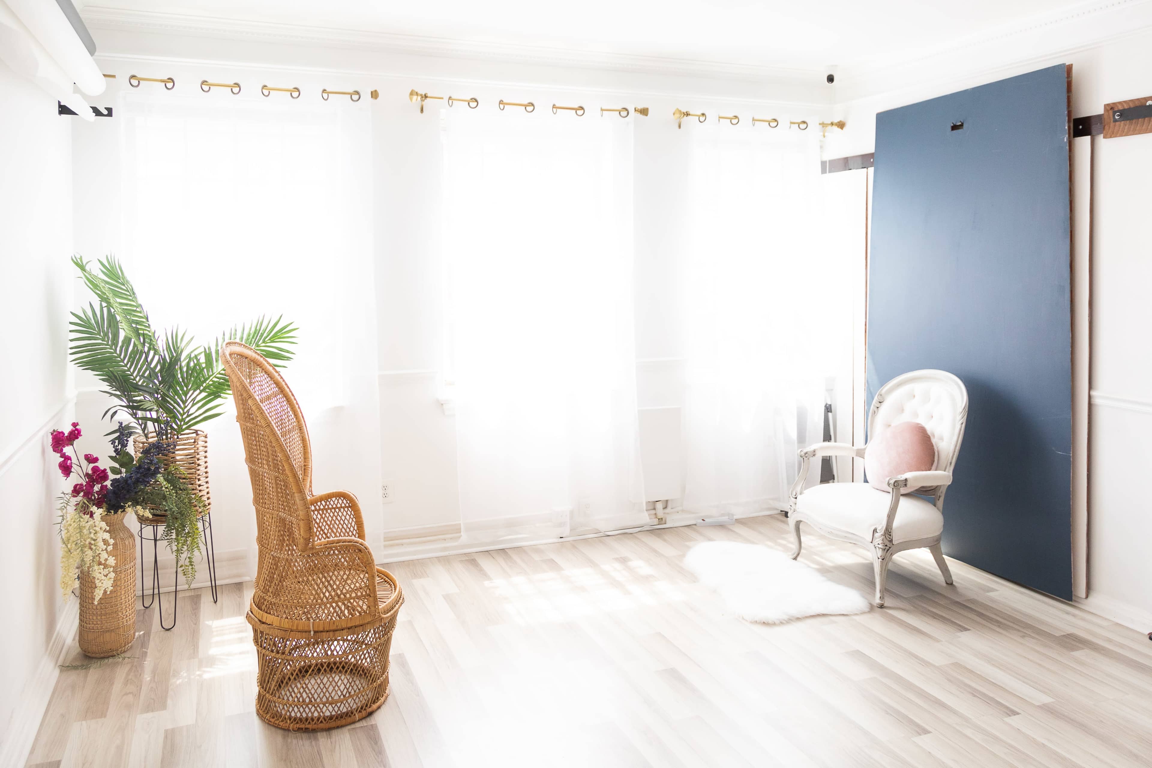 The scene depicts a bright room with large windows, a wicker chair, a white upholstered chair, and a blue wall panel.
