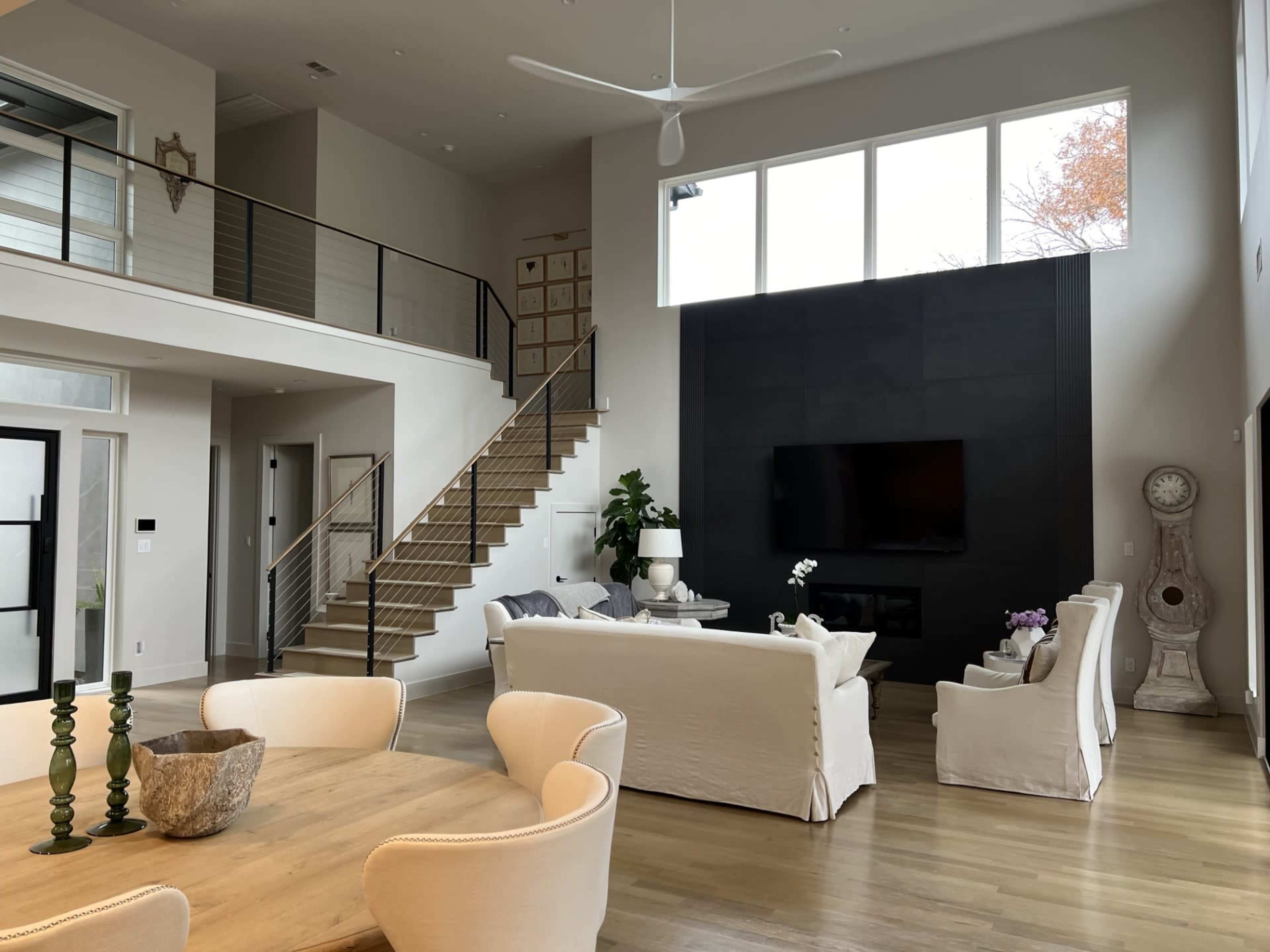 A modern open-concept living room with a staircase, a large wall-mounted TV, and light-colored furniture around a wooden dining table.