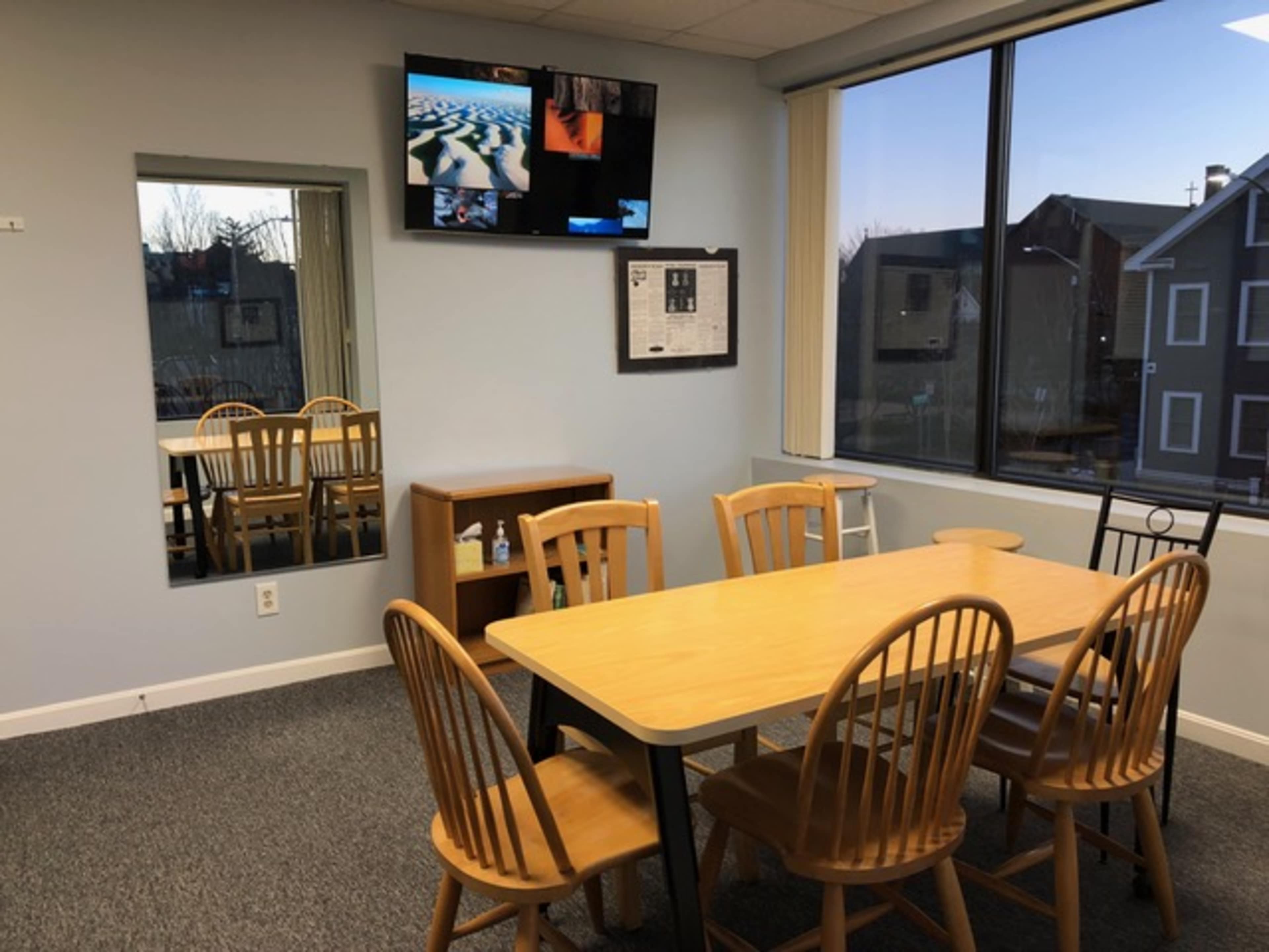 A room features a wooden table surrounded by six chairs, a television mounted on the wall, and a mirror reflecting the space.