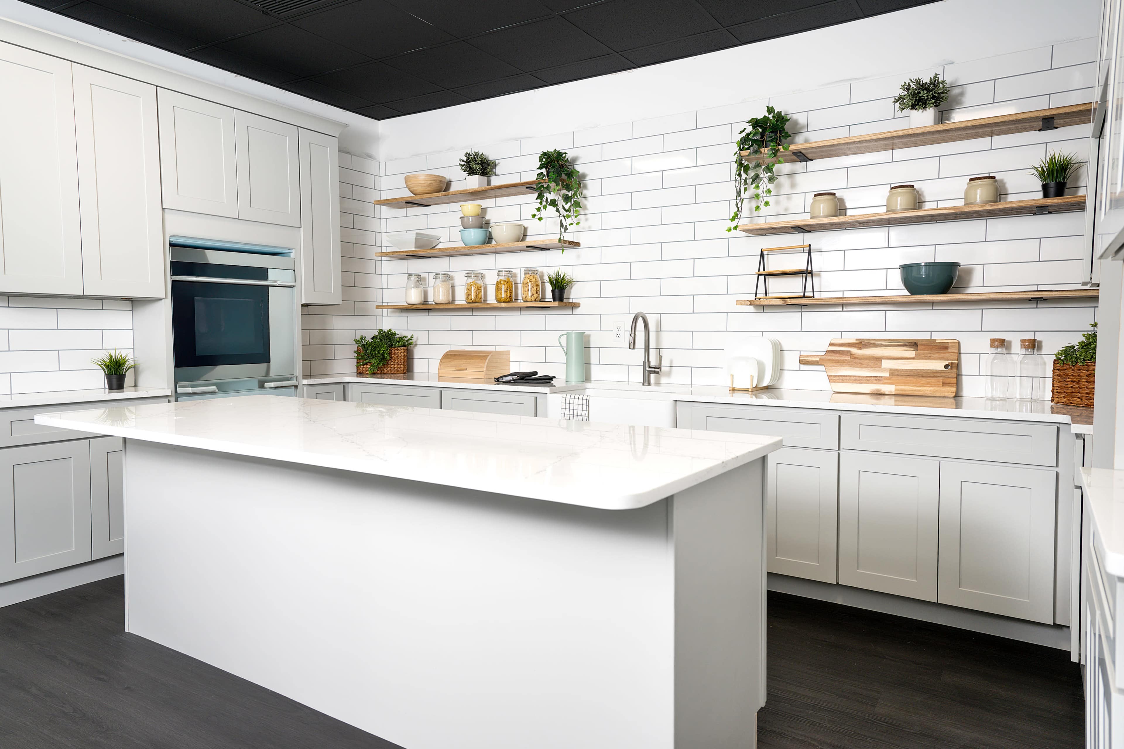 The image shows a modern kitchen with white cabinets, a large island counter, and open shelving displaying various kitchen items and plants.