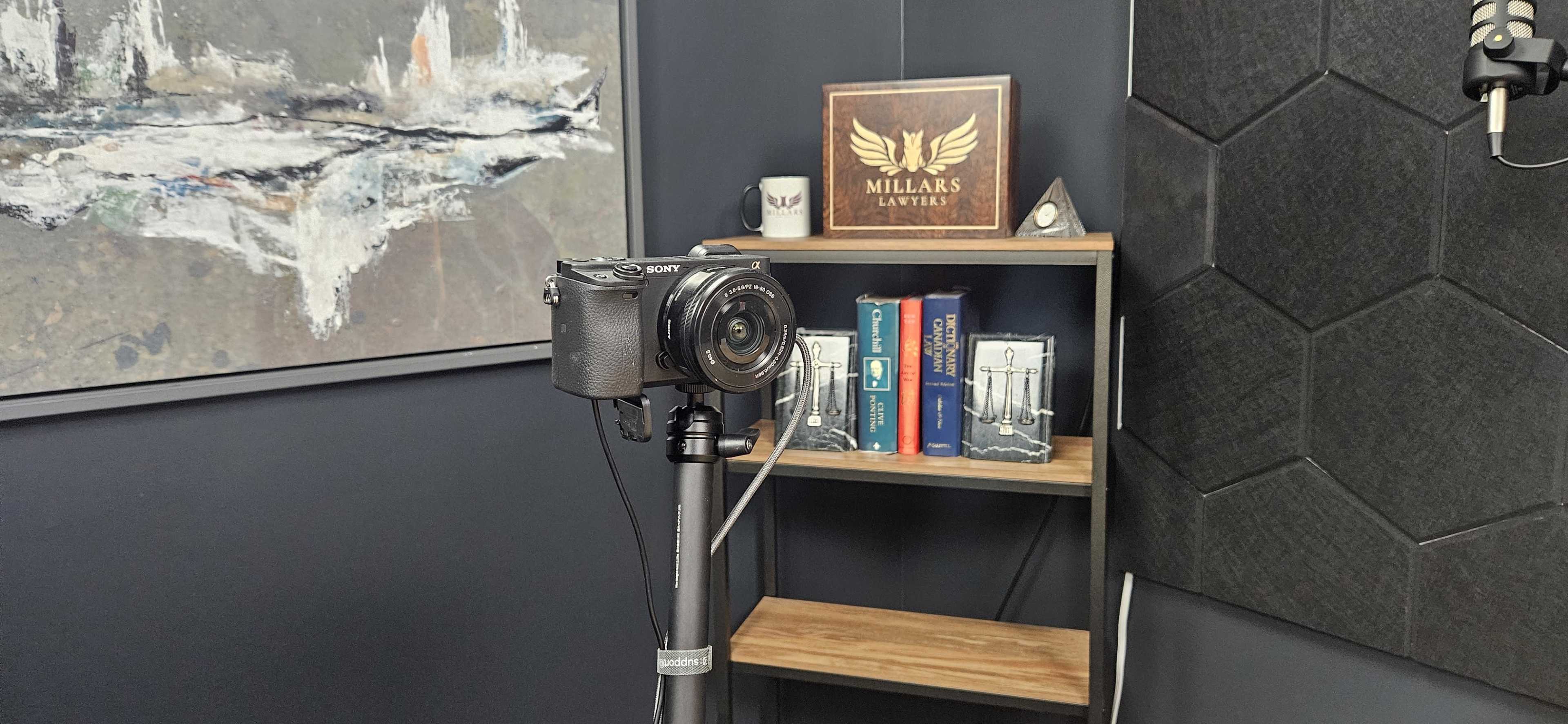 The image shows a black camera mounted on a tripod facing a bookshelf featuring various books and decorative items against a dark wall.