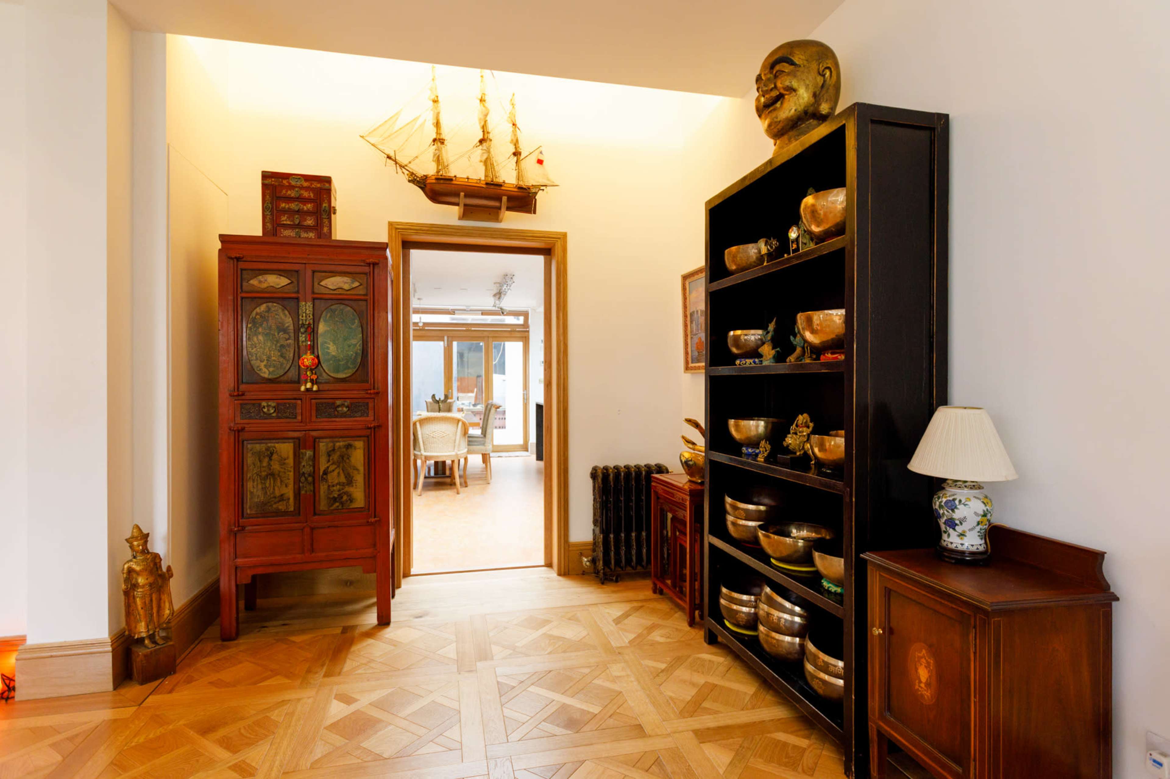 The image shows a hallway with wooden flooring, featuring an ornate cabinet, shelves with metallic bowls, a statue, and a model ship on display.