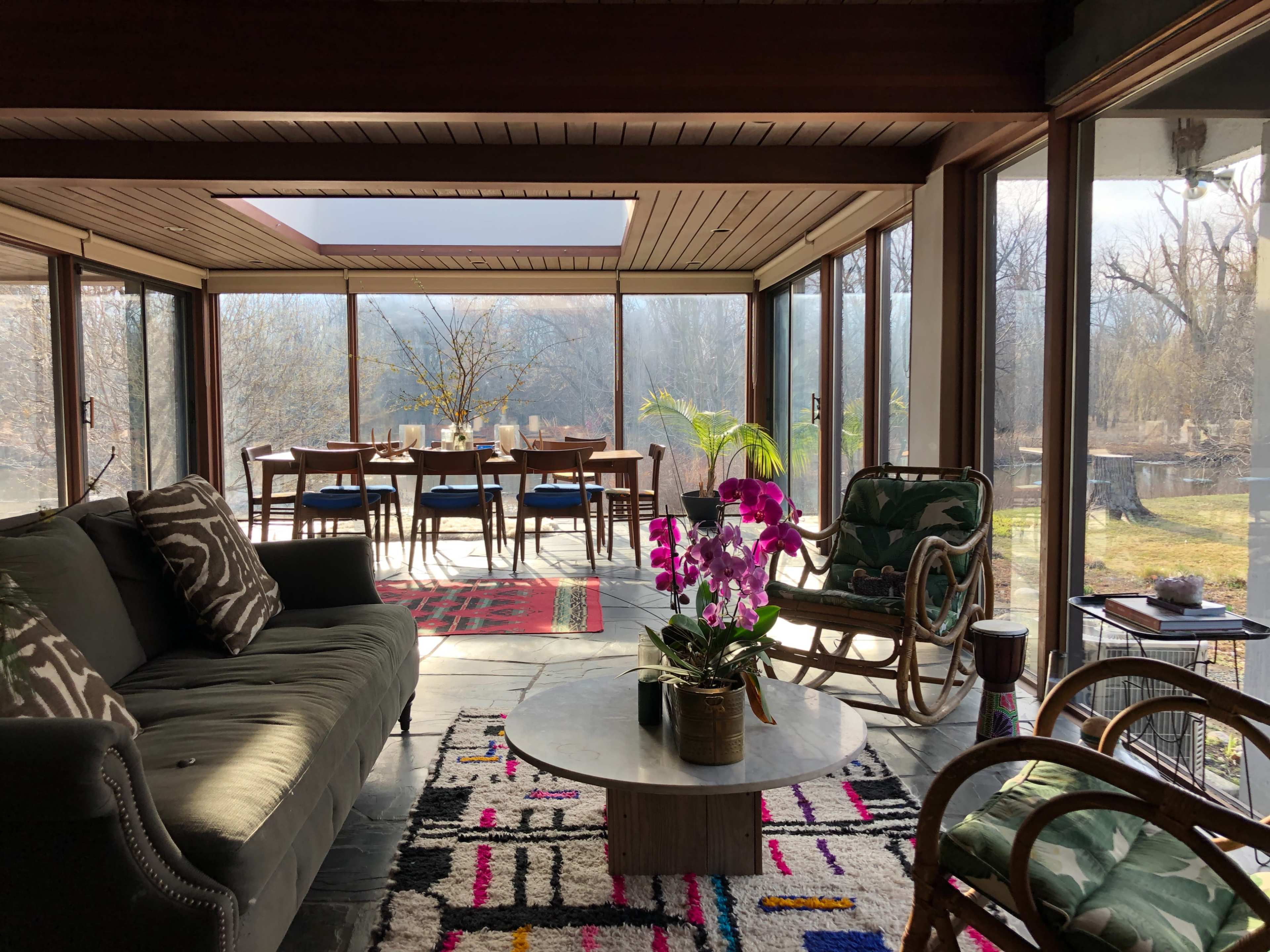 The image shows a modern living space with large glass windows, featuring a couch, a coffee table with a plant, and a dining area set for several people.