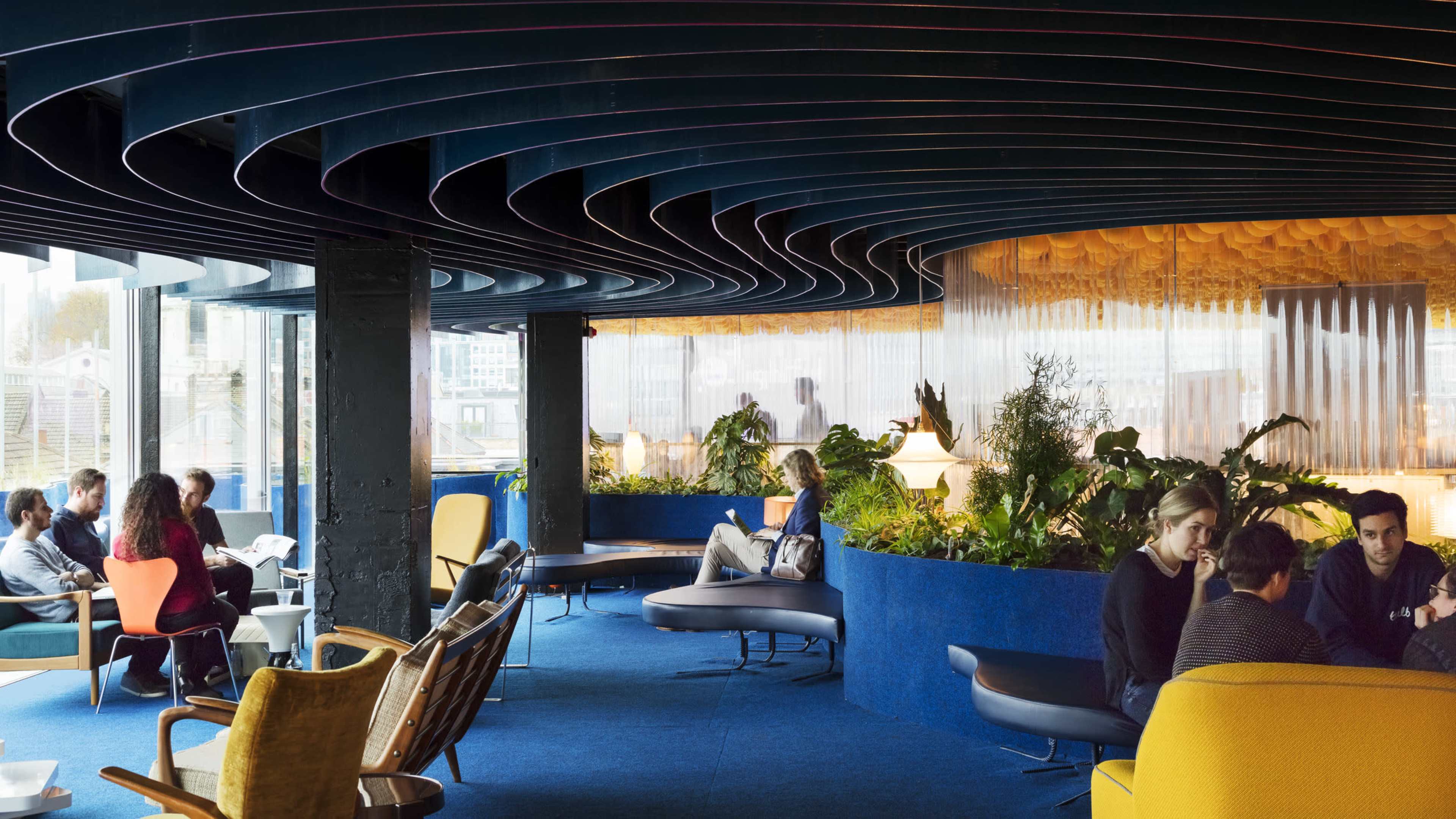 The image showcases a modern interior space featuring curved ceiling design, various seating arrangements, and greenery, with people engaged in conversation and relaxation.