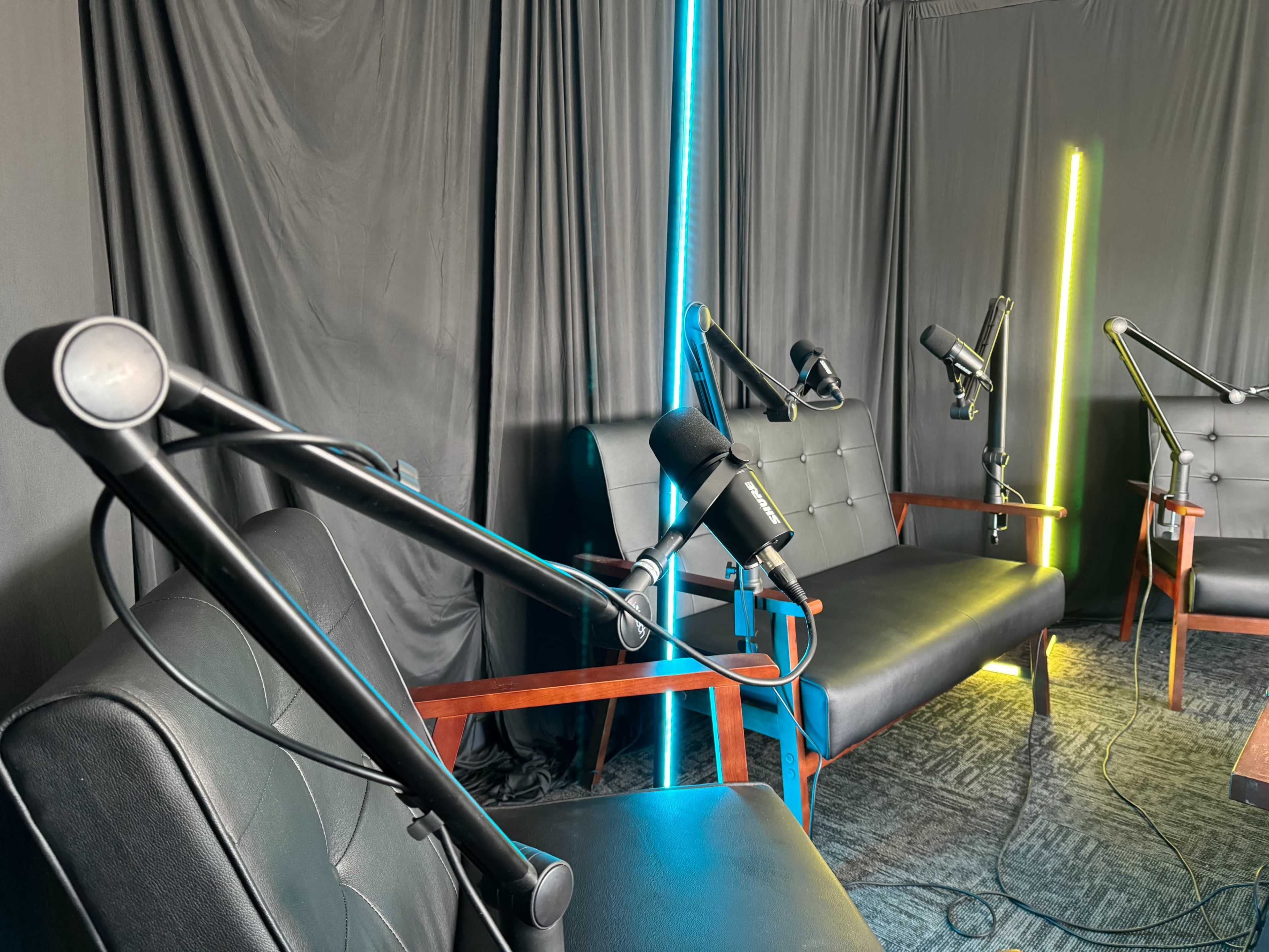 A recording studio with two leather sofas and microphone stands positioned in front of black drapes and illuminated by colored lights.