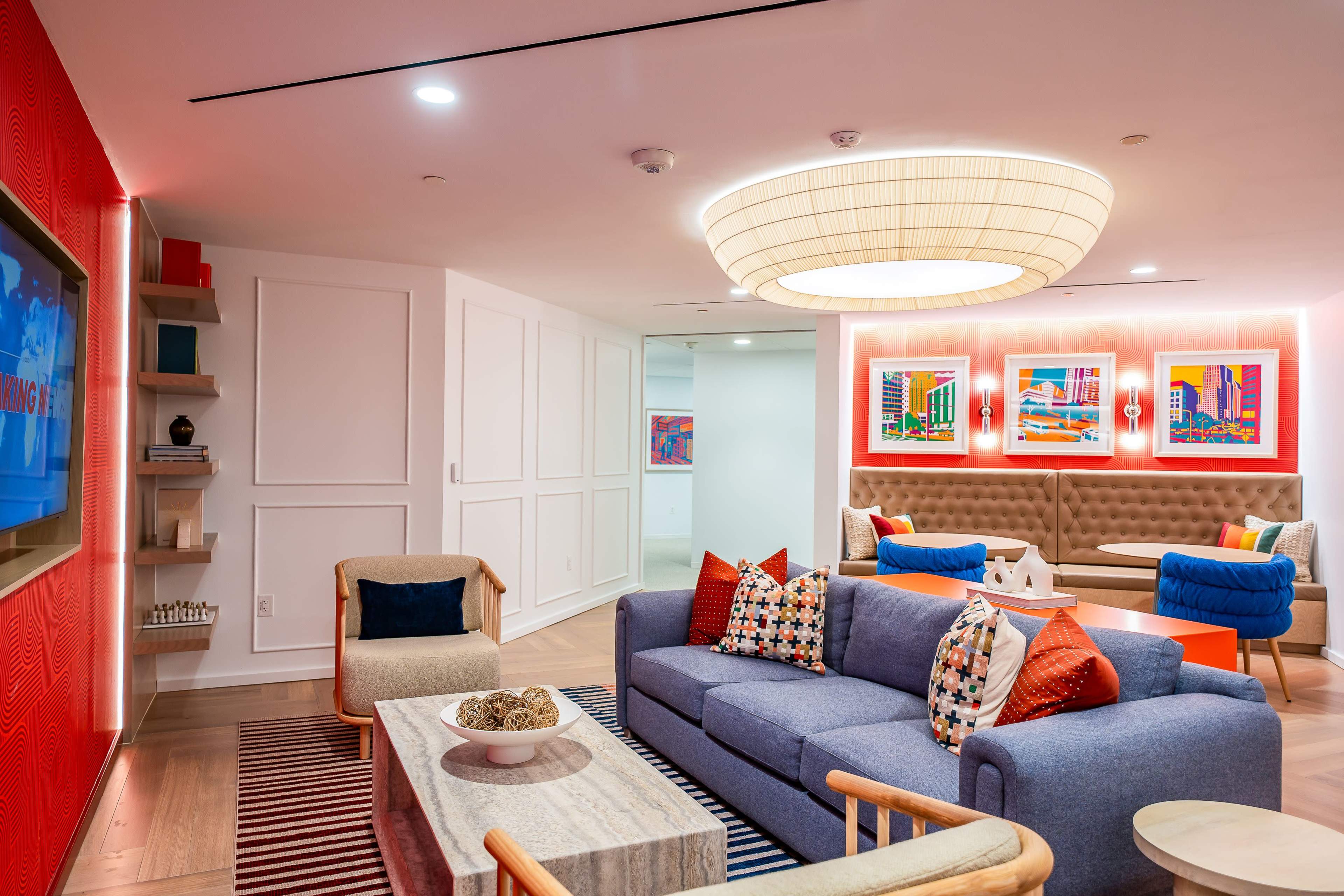 The image shows a modern lounge area with colorful artwork on the walls, a blue sofa with patterned pillows, and a cozy seating arrangement featuring a coffee table and additional chairs.