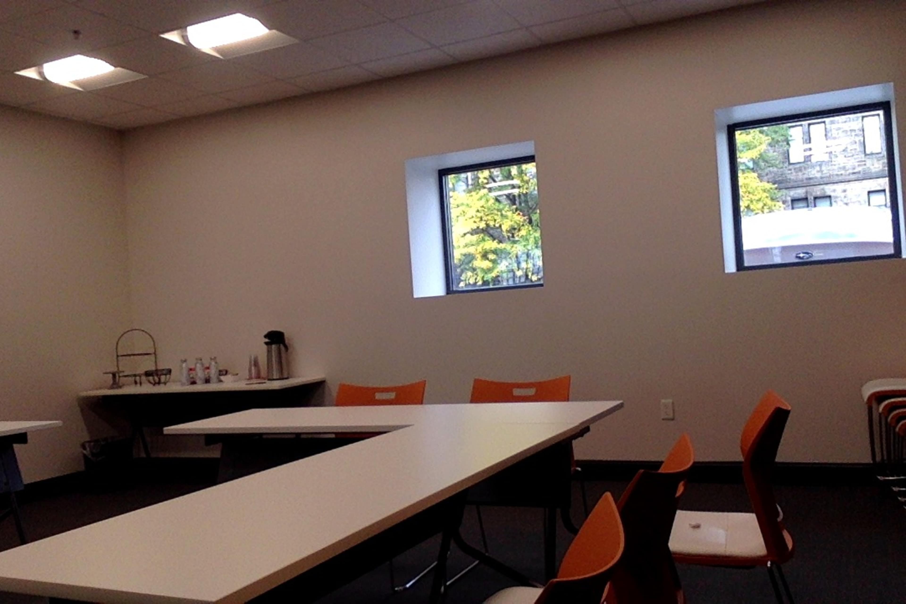 A conference room with two windows, white tables arranged in a U-shape, and orange chairs.