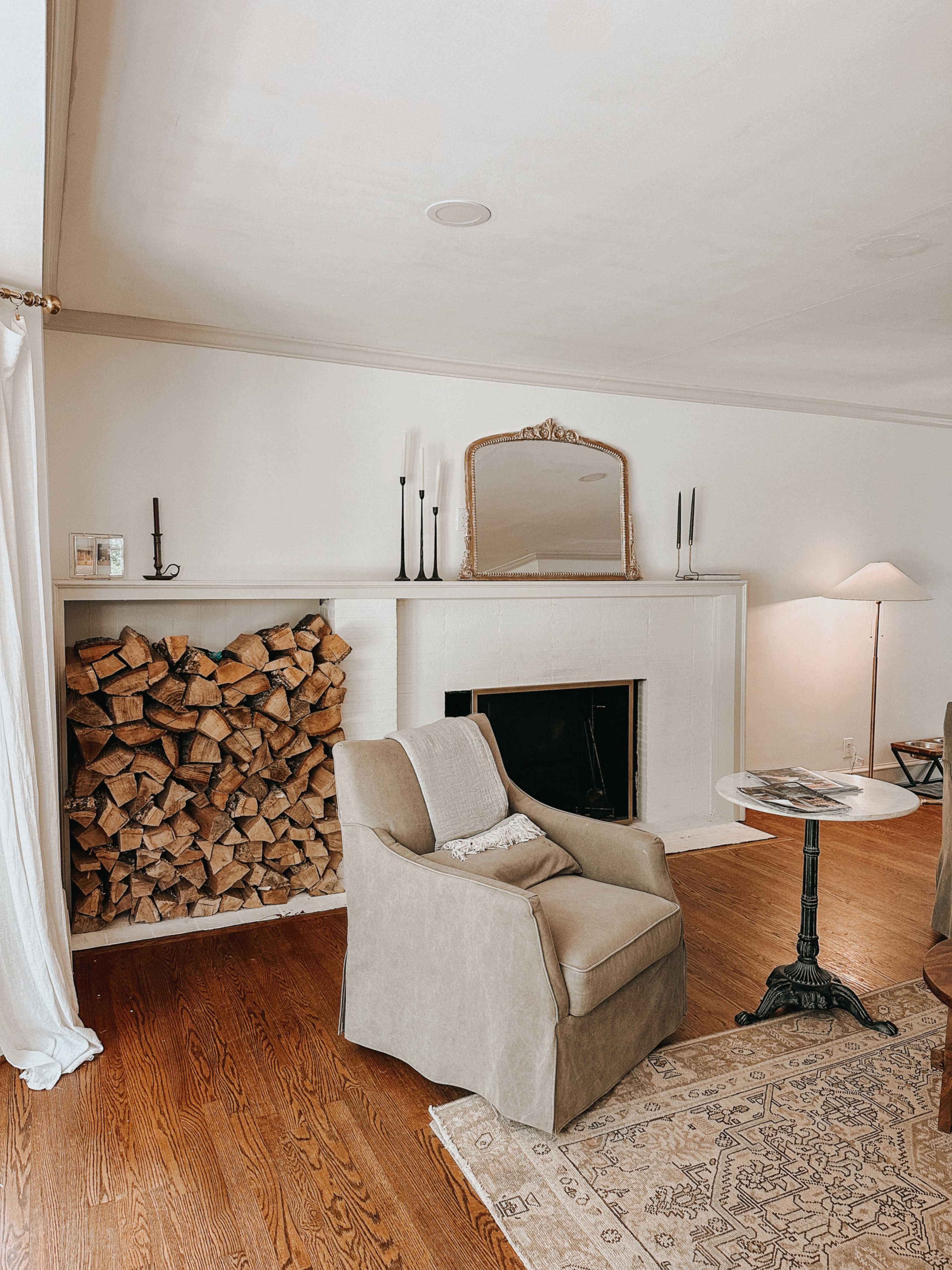 A living room with a fireplace stacked with firewood, a pair of chairs, and a small table beside a lamp.