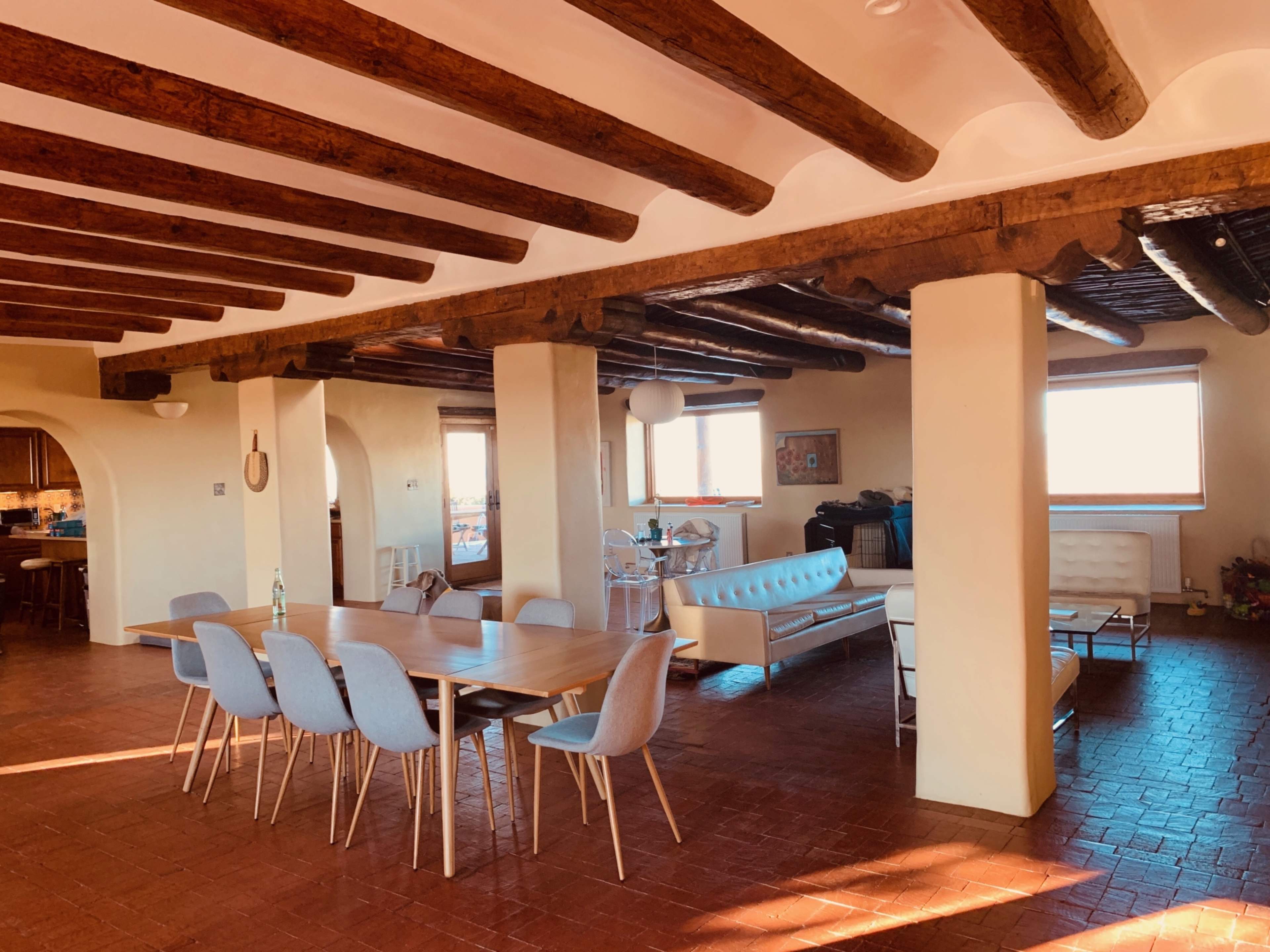 A spacious interior of a house with exposed wooden beams, a long dining table surrounded by light blue chairs, and a white couch in a living area.