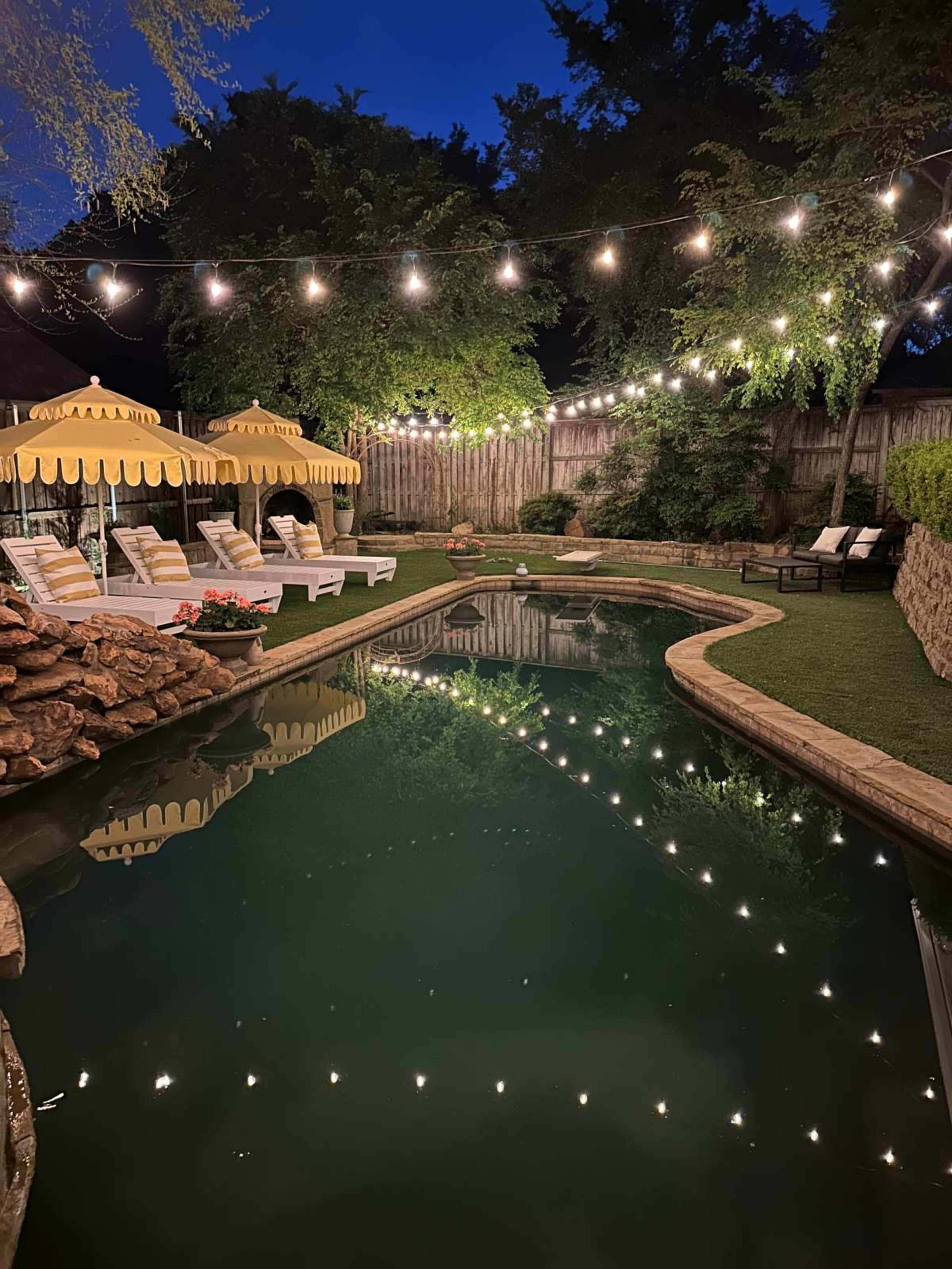 A backyard pool area features lounge chairs and umbrellas, illuminated by string lights along the water's edge.