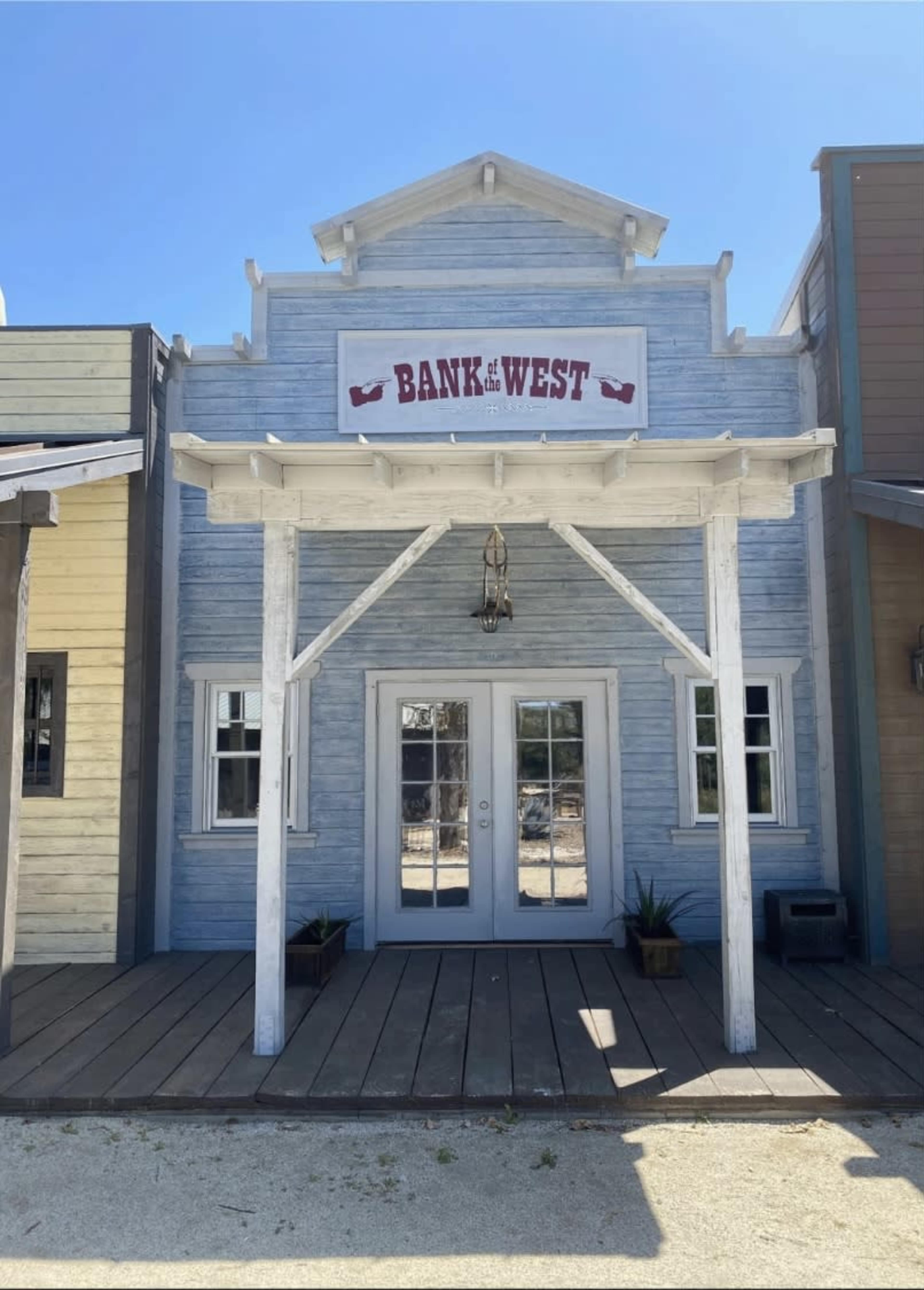 The image shows a western-style bank building with a sign reading "BANK OF THE WEST" above its double doors, framed by a porch and windows on either side.