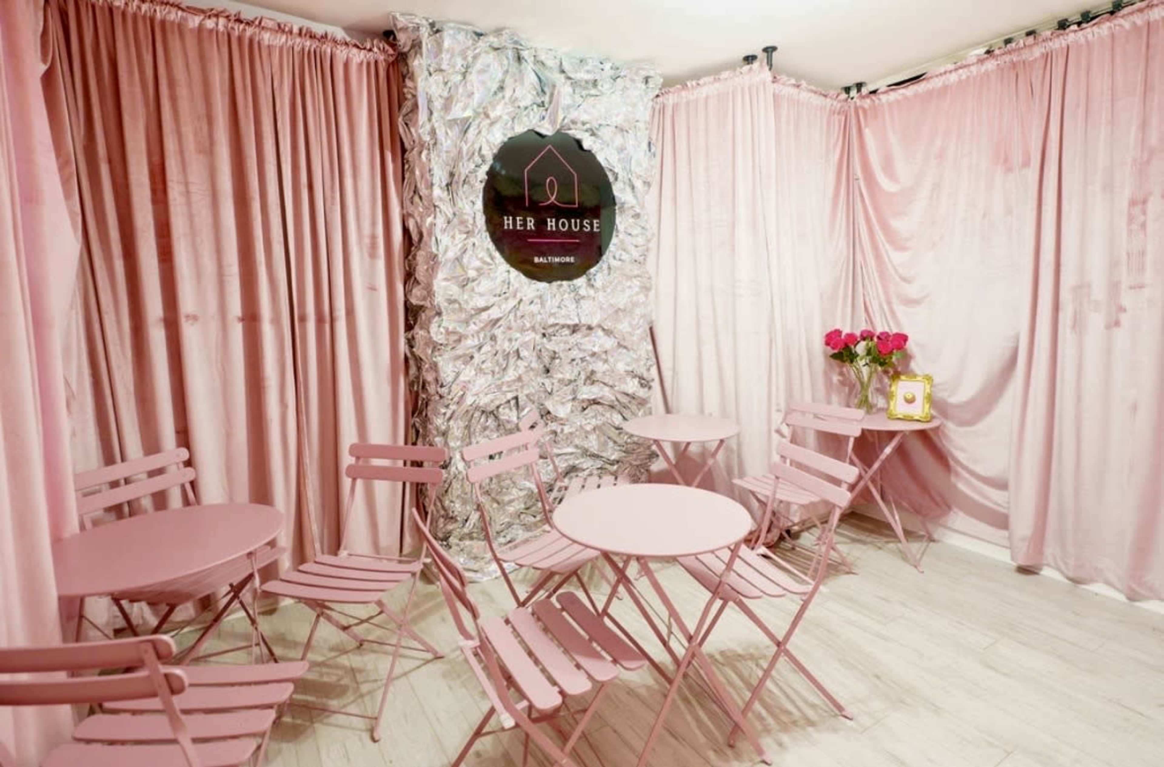 The image shows a small indoor space adorned with pink curtains, featuring two round pink tables with matching chairs and a decorative wall with a circular sign that reads "HER HOUSE."