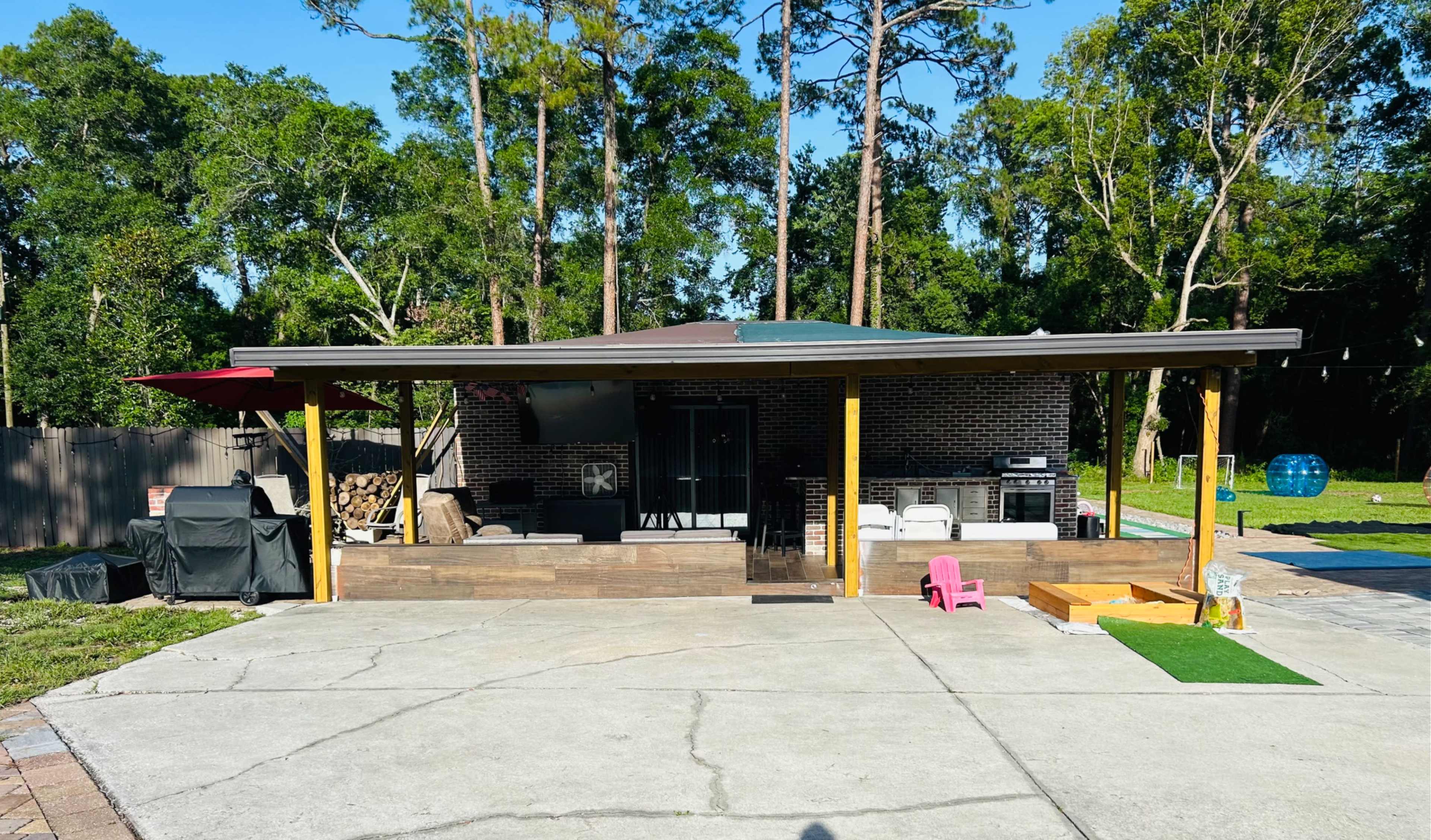 A backyard patio features a brick structure with a covered porch, outdoor furniture, and a barbecue area surrounded by trees.
