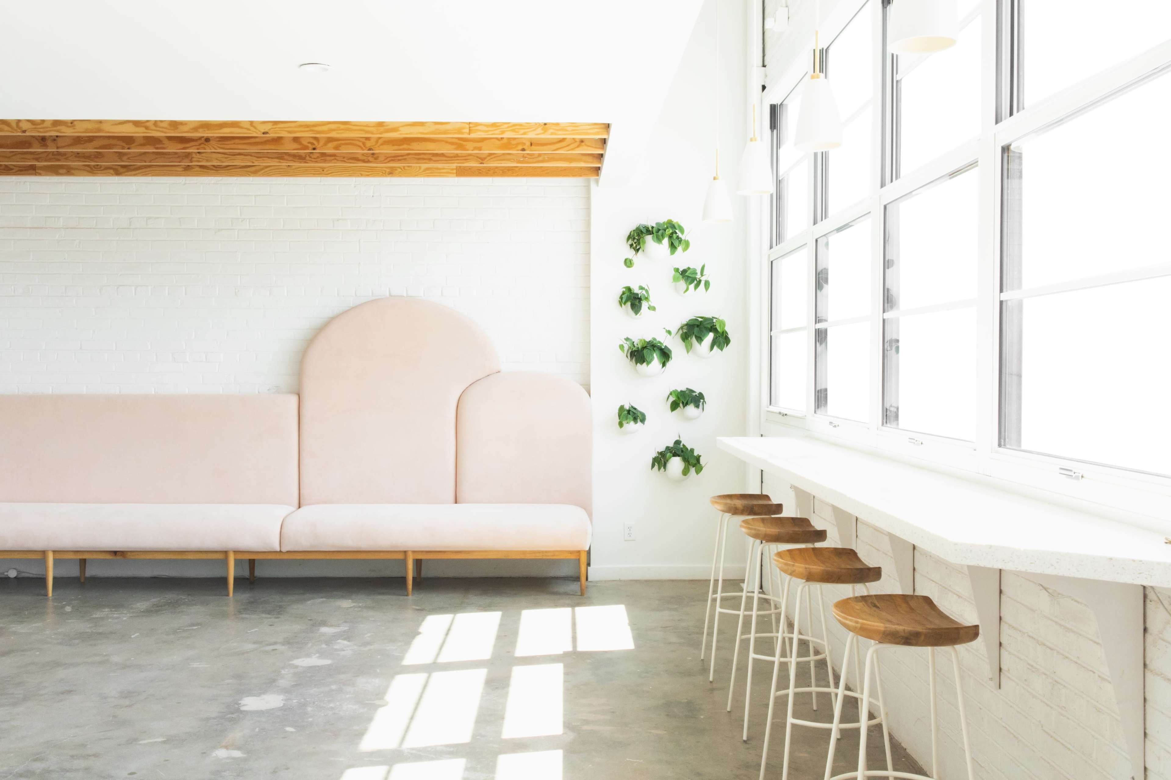 The image shows a bright, minimalist interior featuring a pale pink sofa, a row of wooden bar stools, and green plants against a white brick wall.