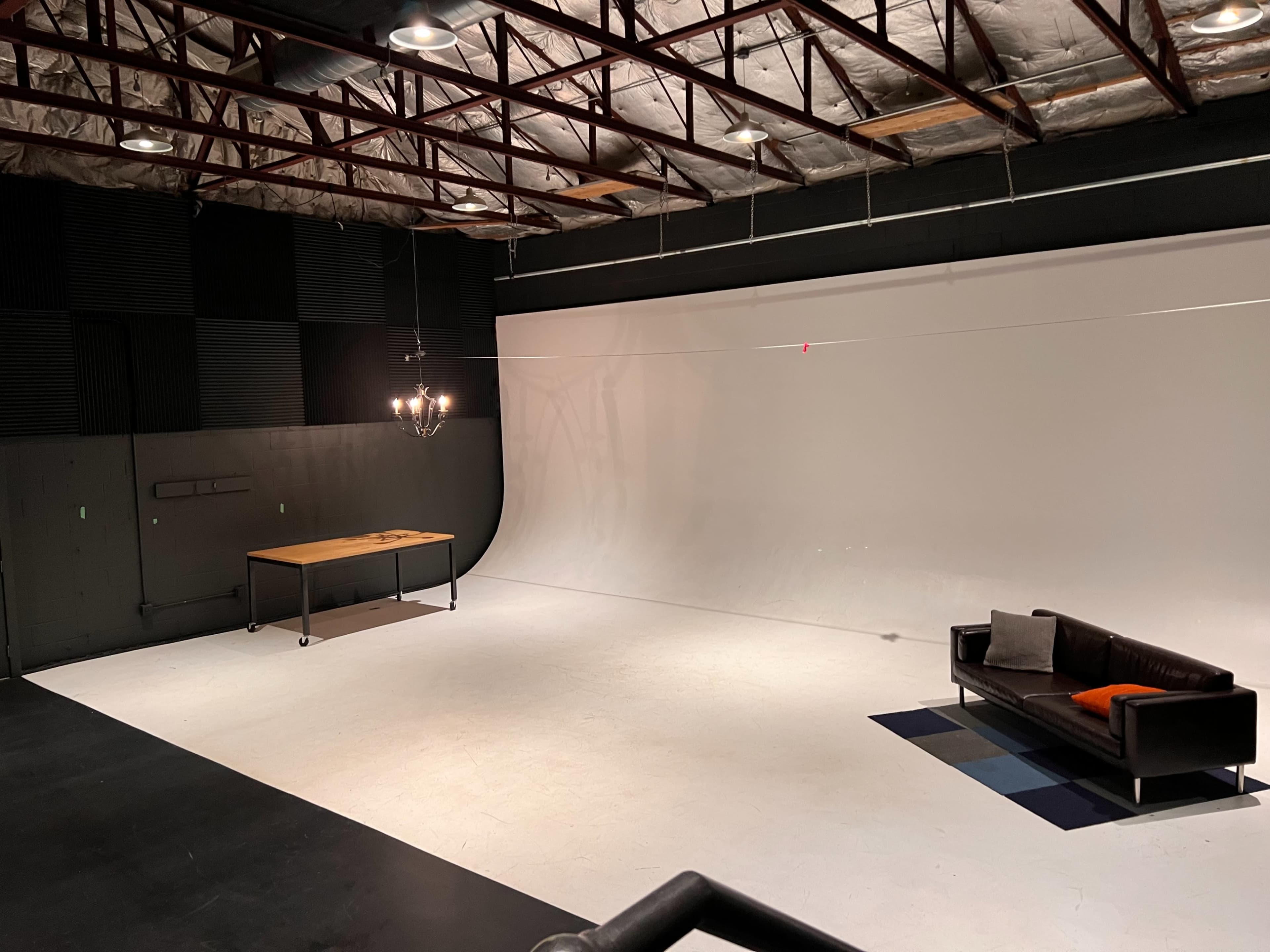 A spacious studio with a white backdrop, a wooden table, a black sofa, and a patterned rug on the floor.
