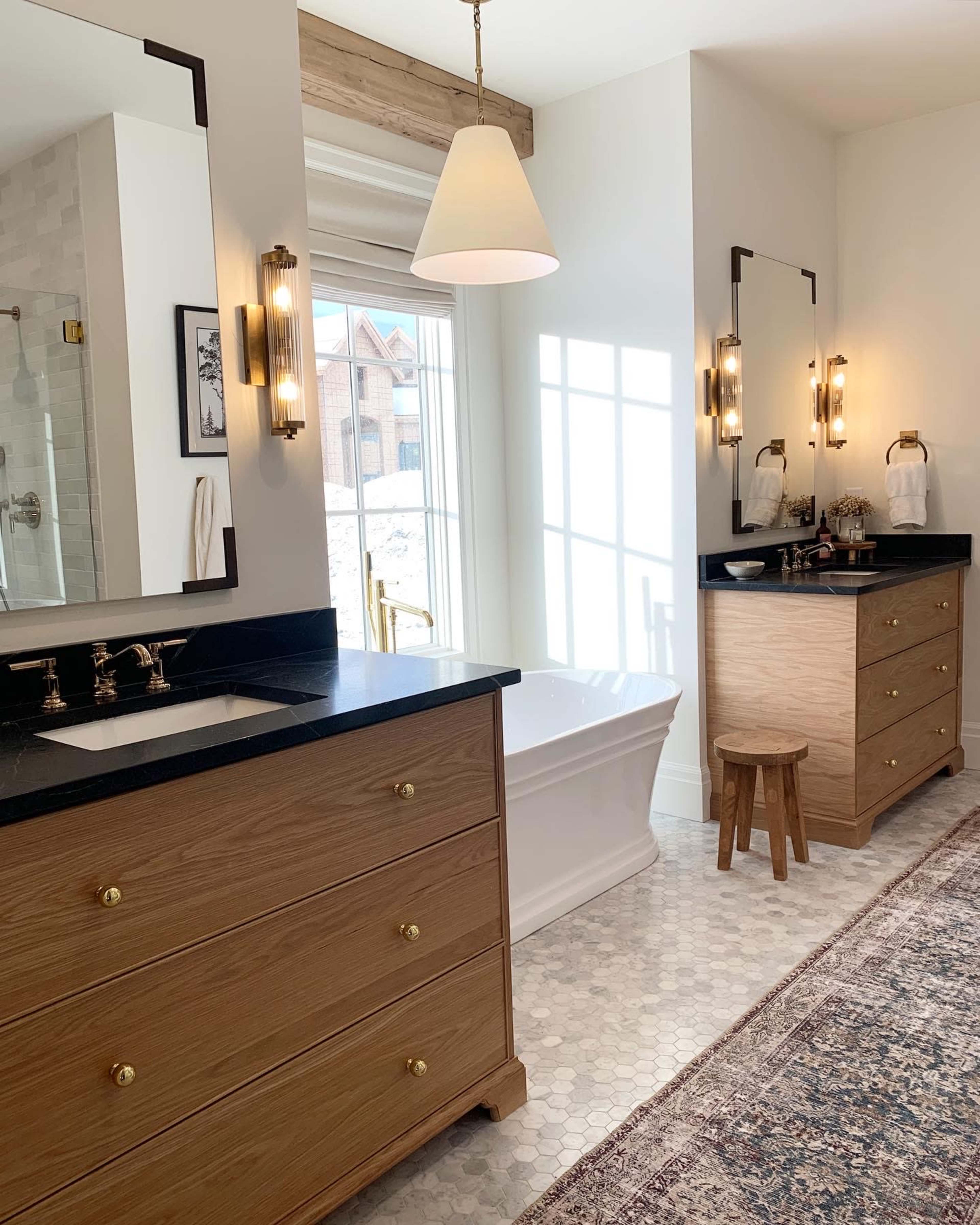 A bright bathroom featuring a freestanding tub, double sinks with a dark countertop, wooden cabinetry, and a window providing natural light.