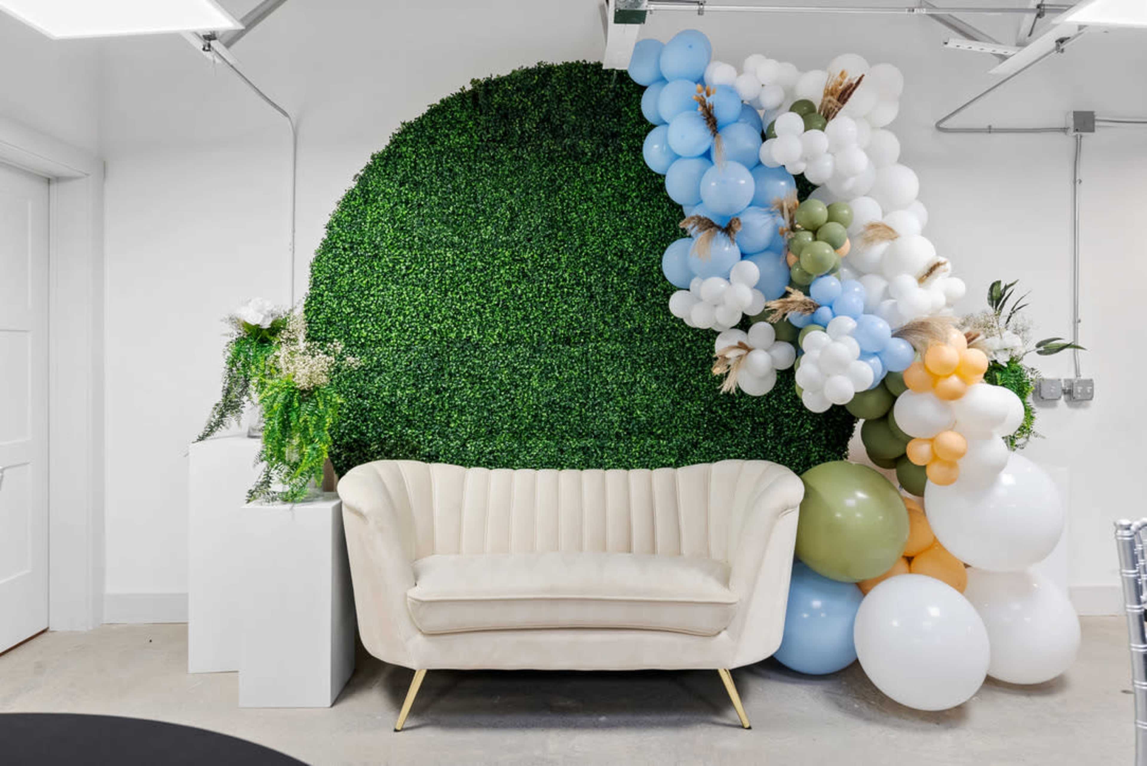 A cream-colored sofa is positioned in front of a green, leafy wall, adorned with an arrangement of colorful balloons.