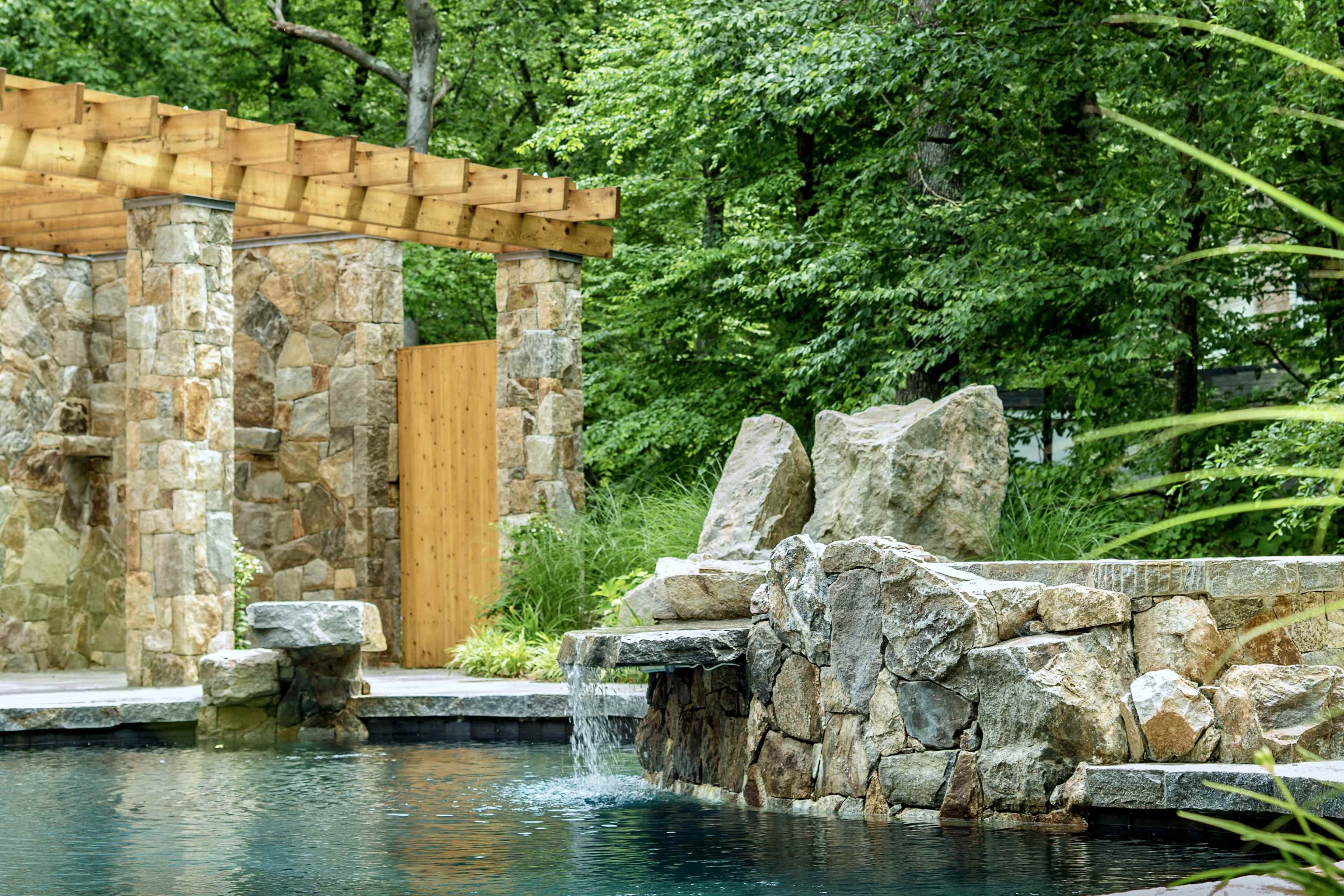 A stone wall surrounds a serene swimming pool with a natural rock feature and a wooden gate in a lush green setting.