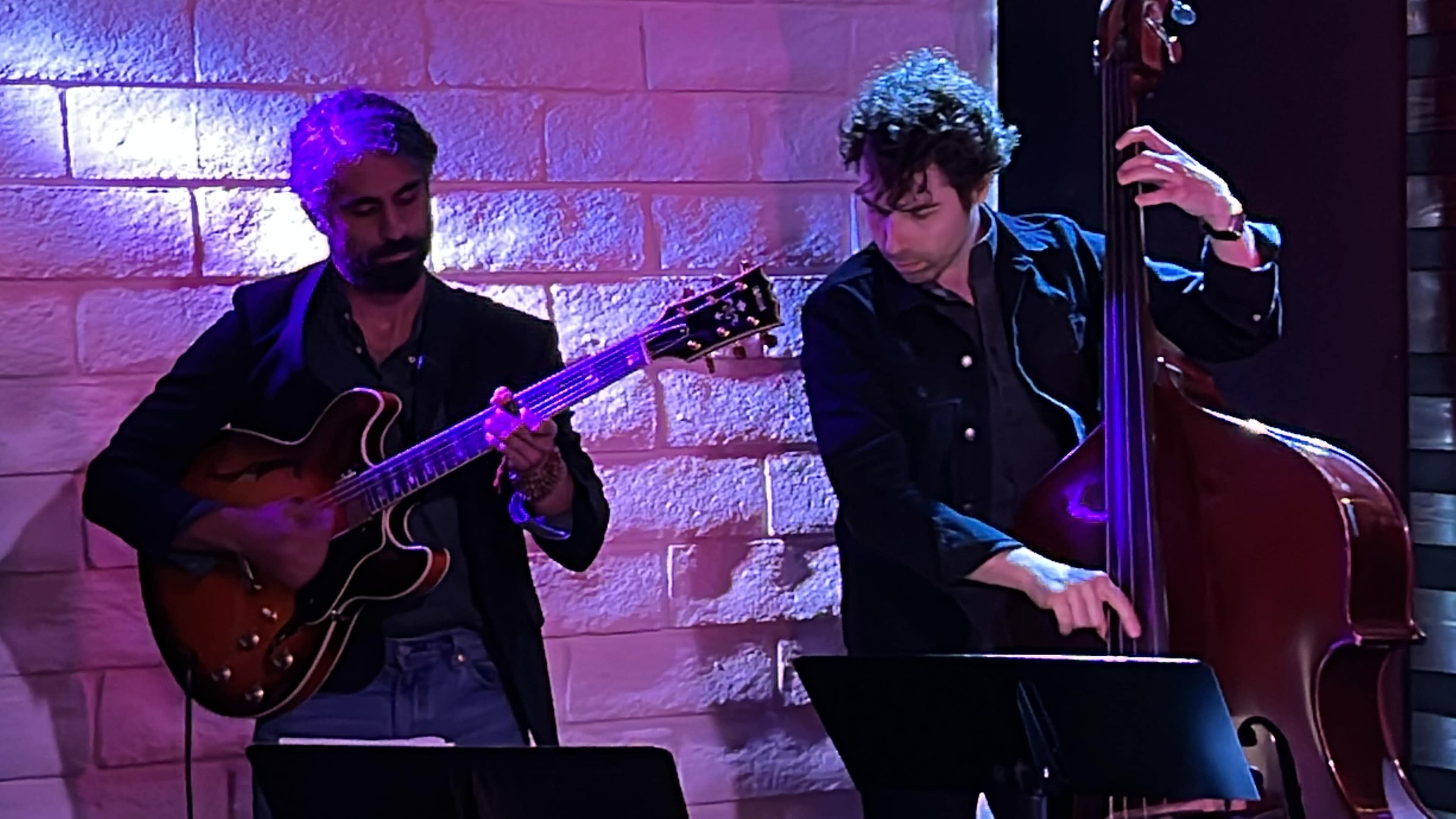 Two musicians perform live on stage, one playing an electric guitar and the other a double bass, against a backdrop of illuminated brick walls.
