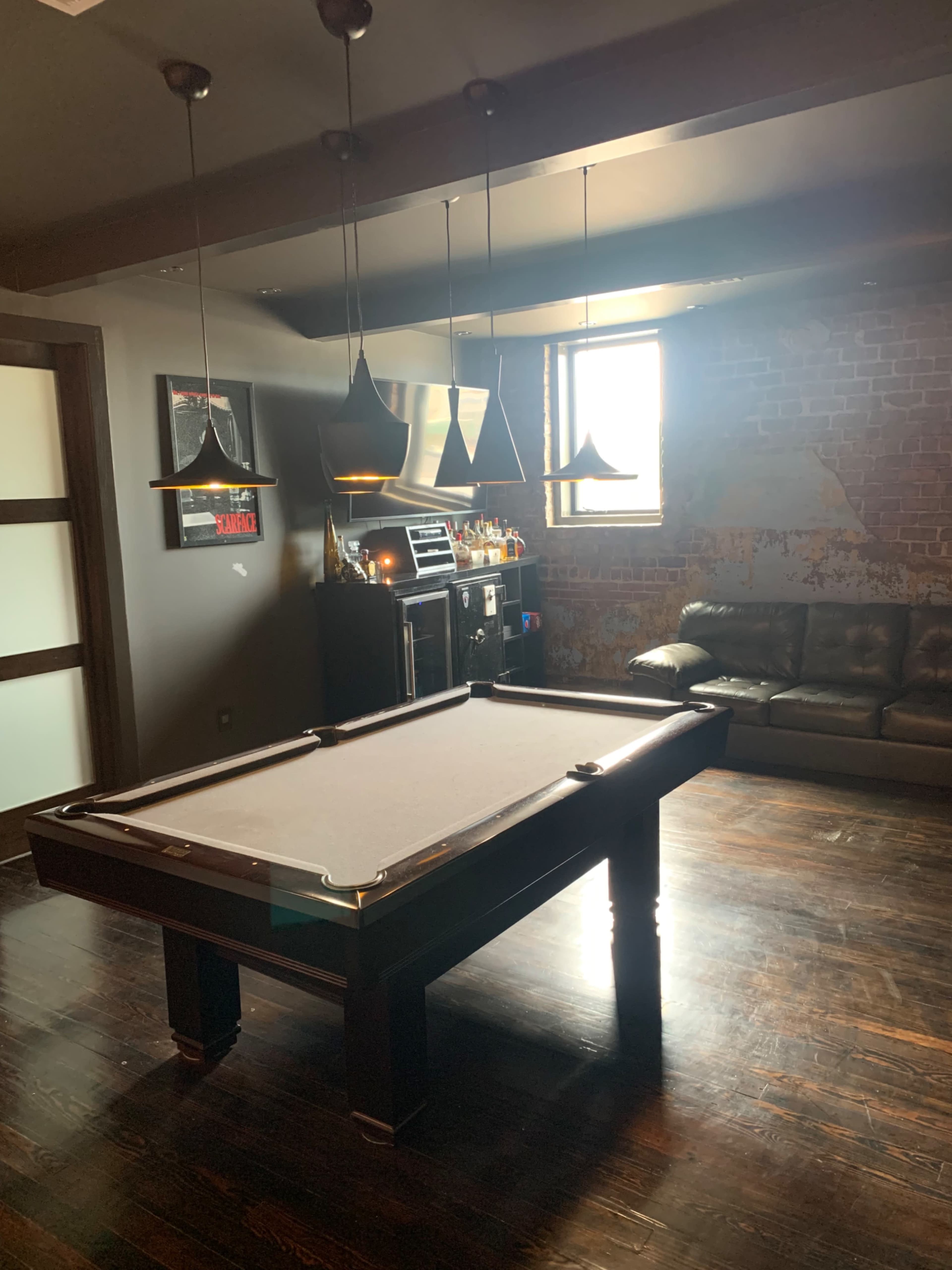 The image shows a billiards table in a spacious room with exposed brick walls, a leather couch, and overhead pendant lights.