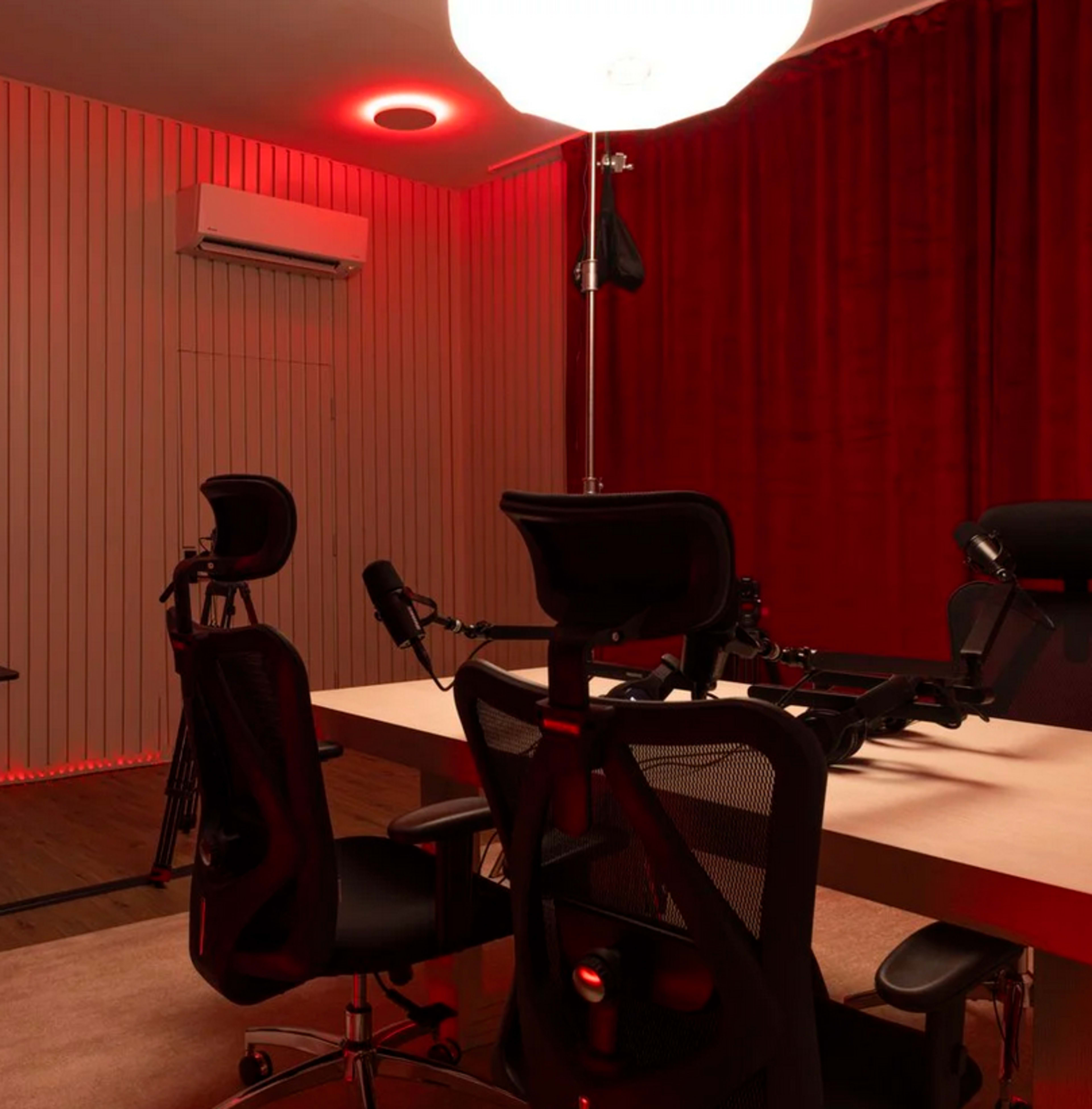 The image shows a modern recording studio with a large table, several black office chairs, and red lighting along the walls and ceiling.