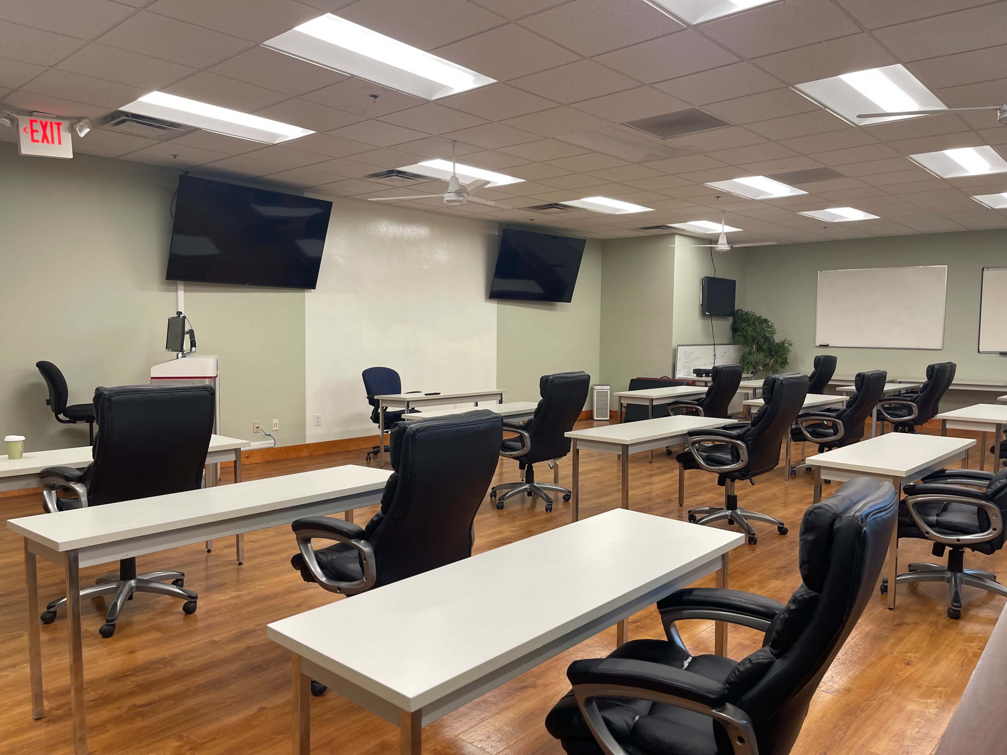 The image shows a classroom setup with several empty desks and ergonomic chairs arranged in a spacious room, equipped with two wall-mounted screens and a whiteboard.