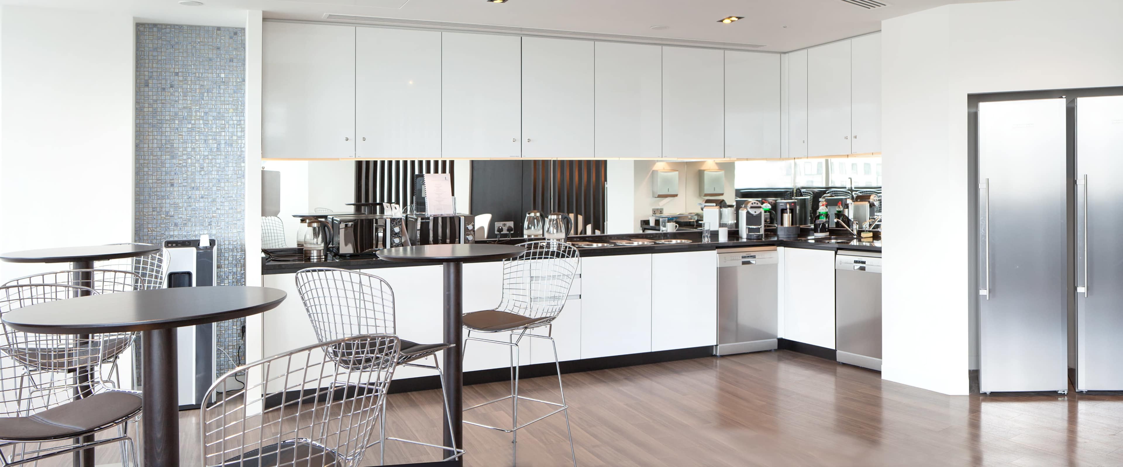 The image shows a modern kitchen with white cabinetry, stainless steel appliances, and several bar stools around black tables.