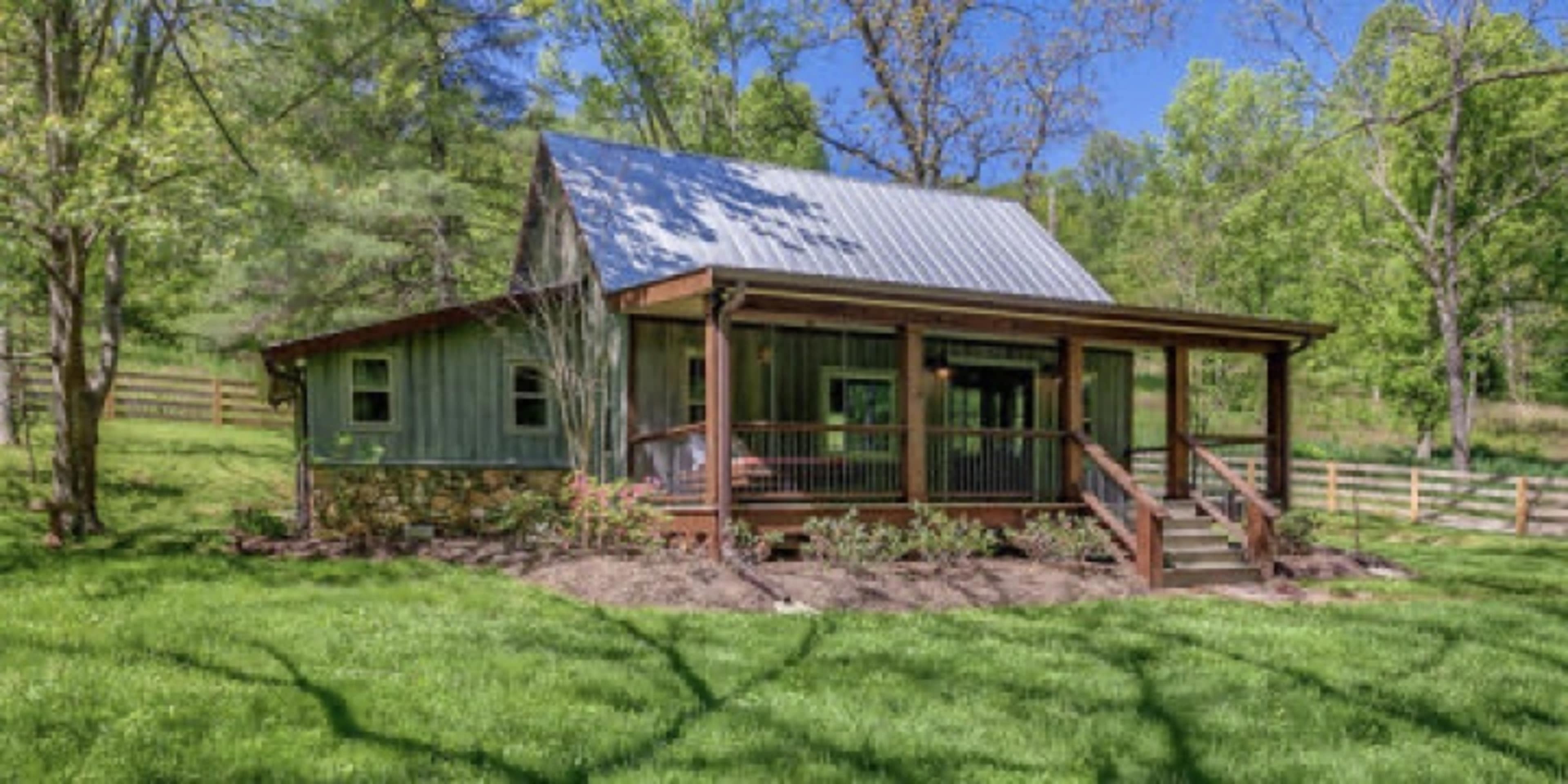 A rustic house with a metal roof and a wooden porch is surrounded by lush green grass and trees.