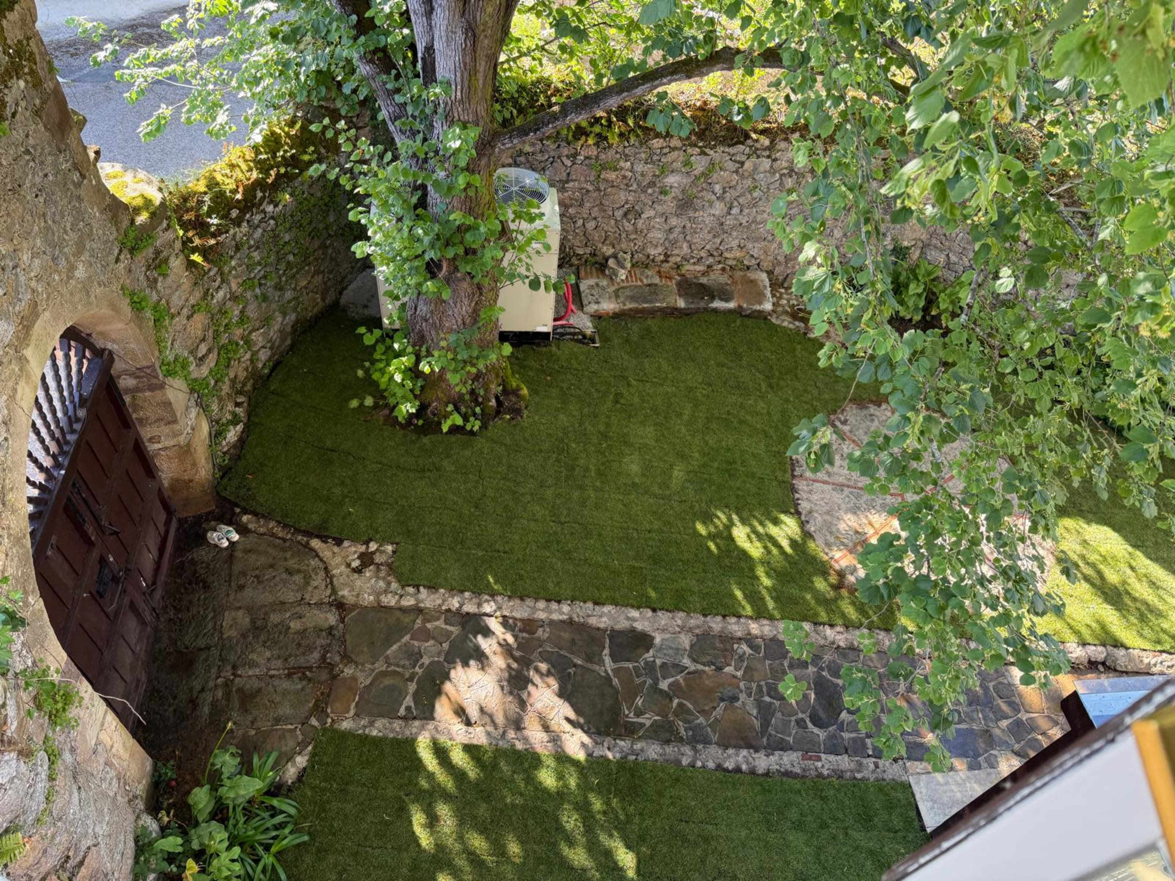 The image shows a landscaped garden area with a stone path, artificial grass, and a large tree, enclosed by a stone wall and wooden gate.