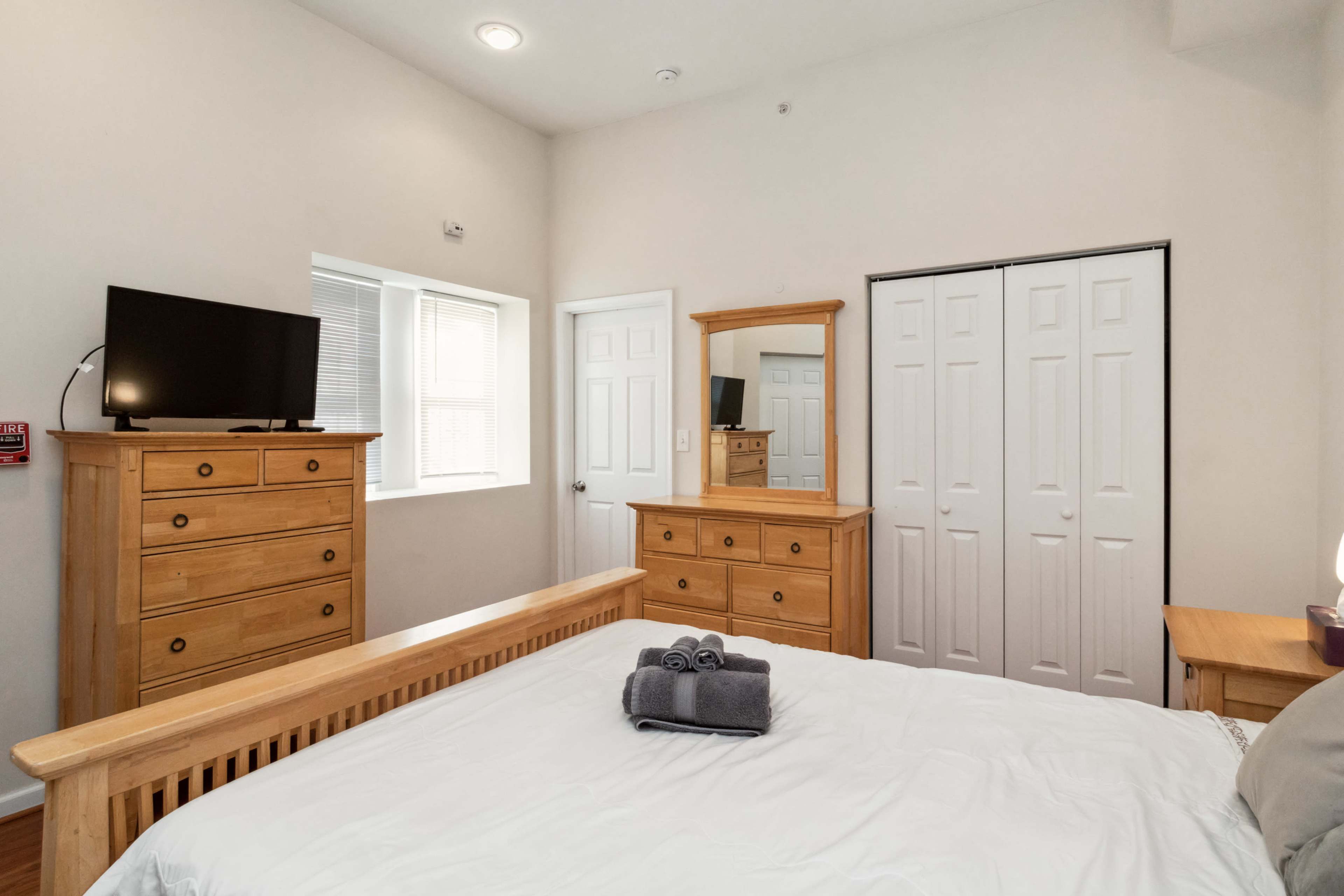 A well-furnished bedroom features a wooden bed, a dresser, a mirrored cabinet, a television, and a window.
