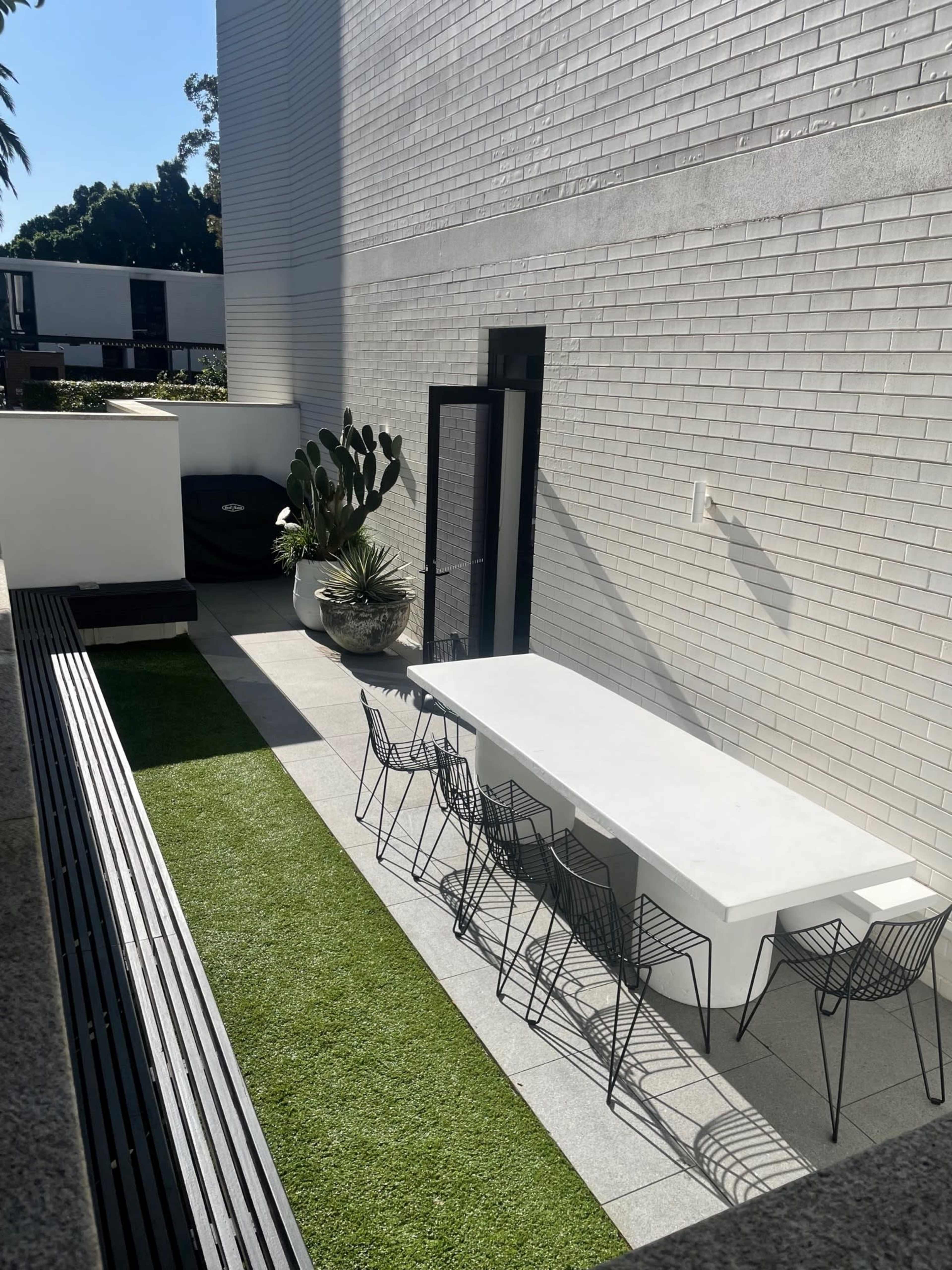 The image shows a narrow outdoor space with a long white dining table and black wire chairs set on green grass beside a wall of white bricks.