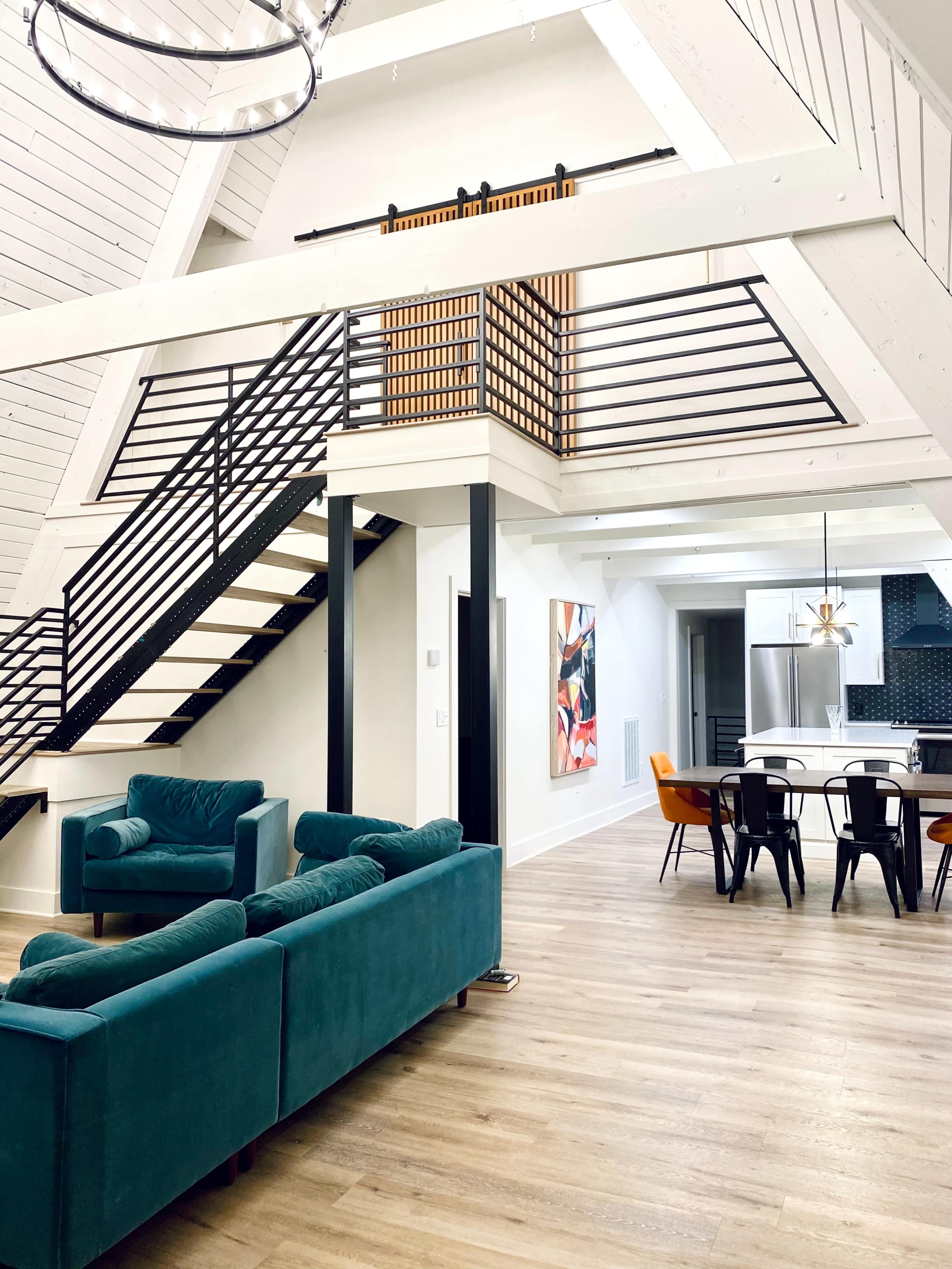 A modern interior with a spacious open layout, featuring a living area with teal sofas, a staircase leading to an upper level, and a dining set adjacent to a kitchen.