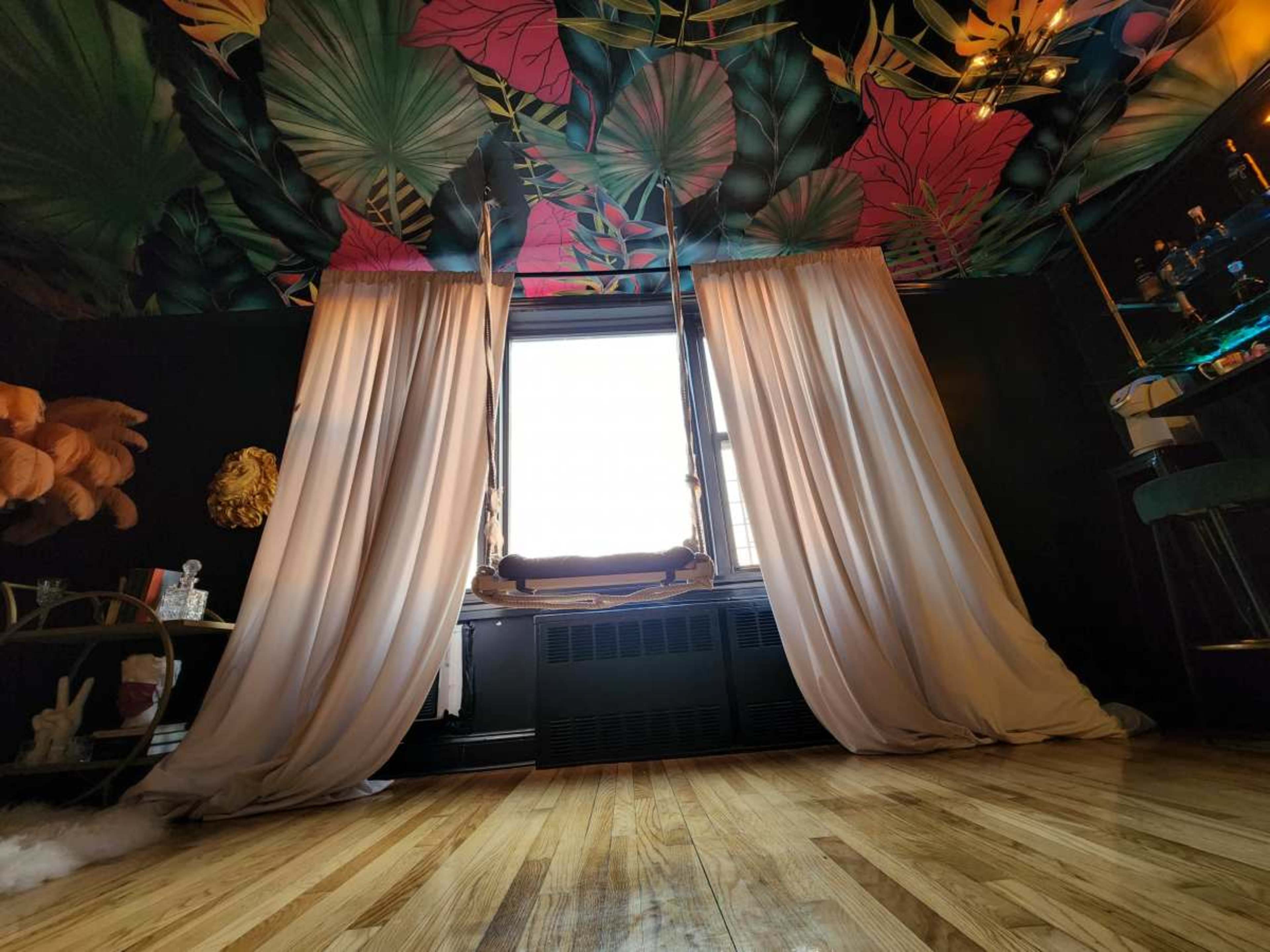 The image shows a room with a colorful floral ceiling, long curtains framing a large window, and wooden flooring.