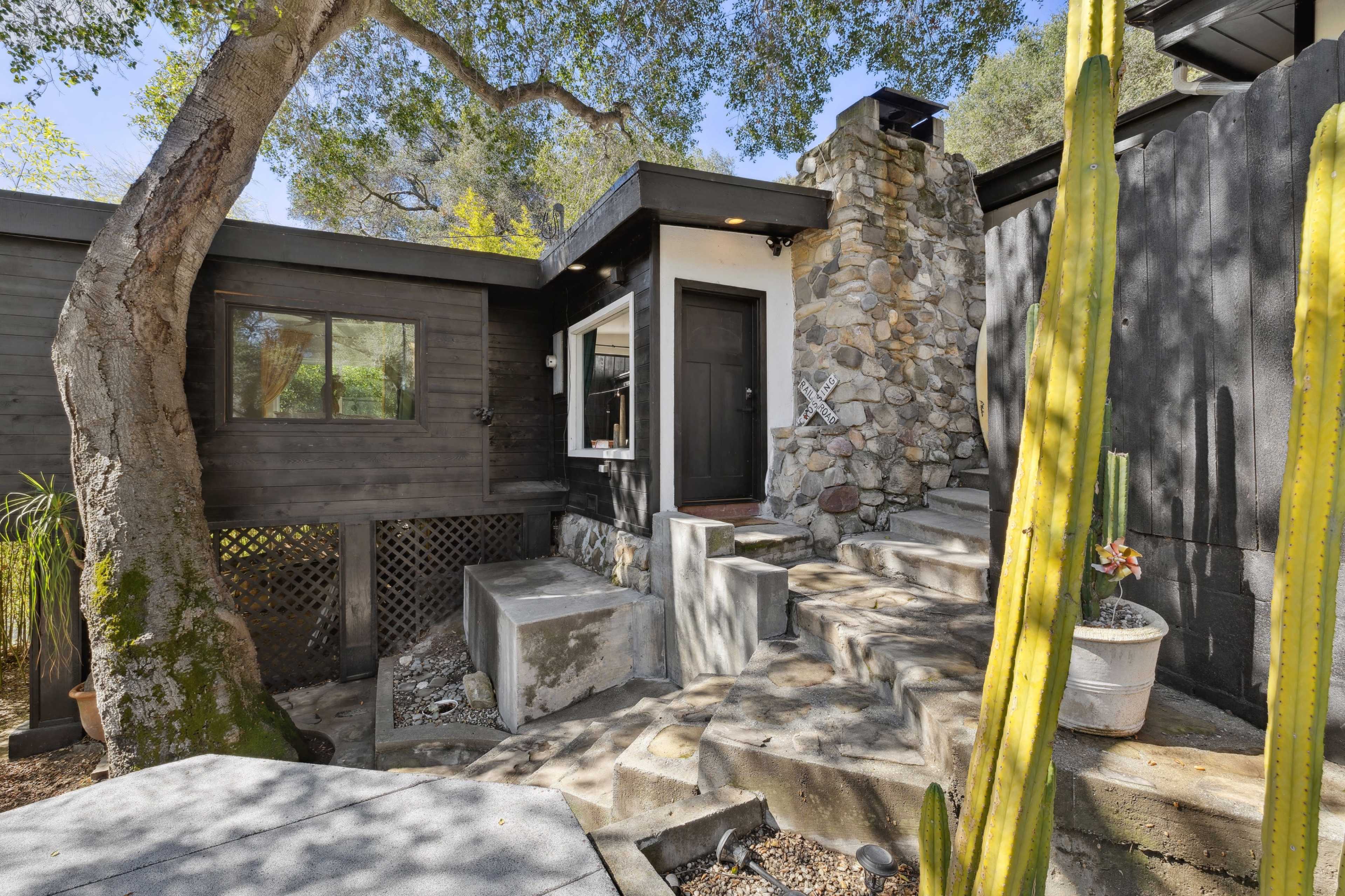 A modern house features a stone chimney and steps leading to a front door, surrounded by greenery and cacti.
