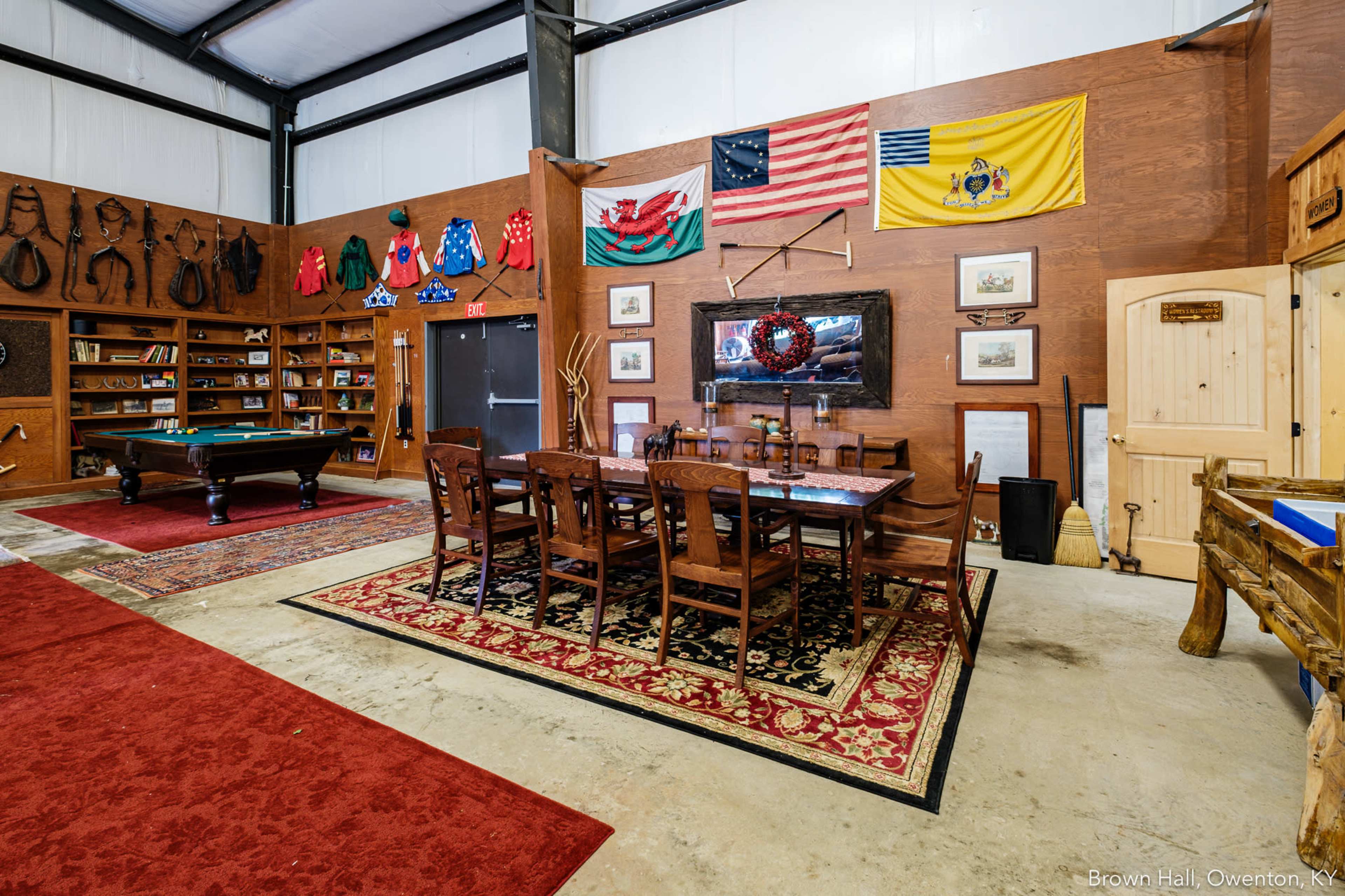 The image shows a rustic dining area within a barn-like structure, featuring a large wooden table surrounded by chairs, pool table, and various decorative flags and exhibits on the walls.