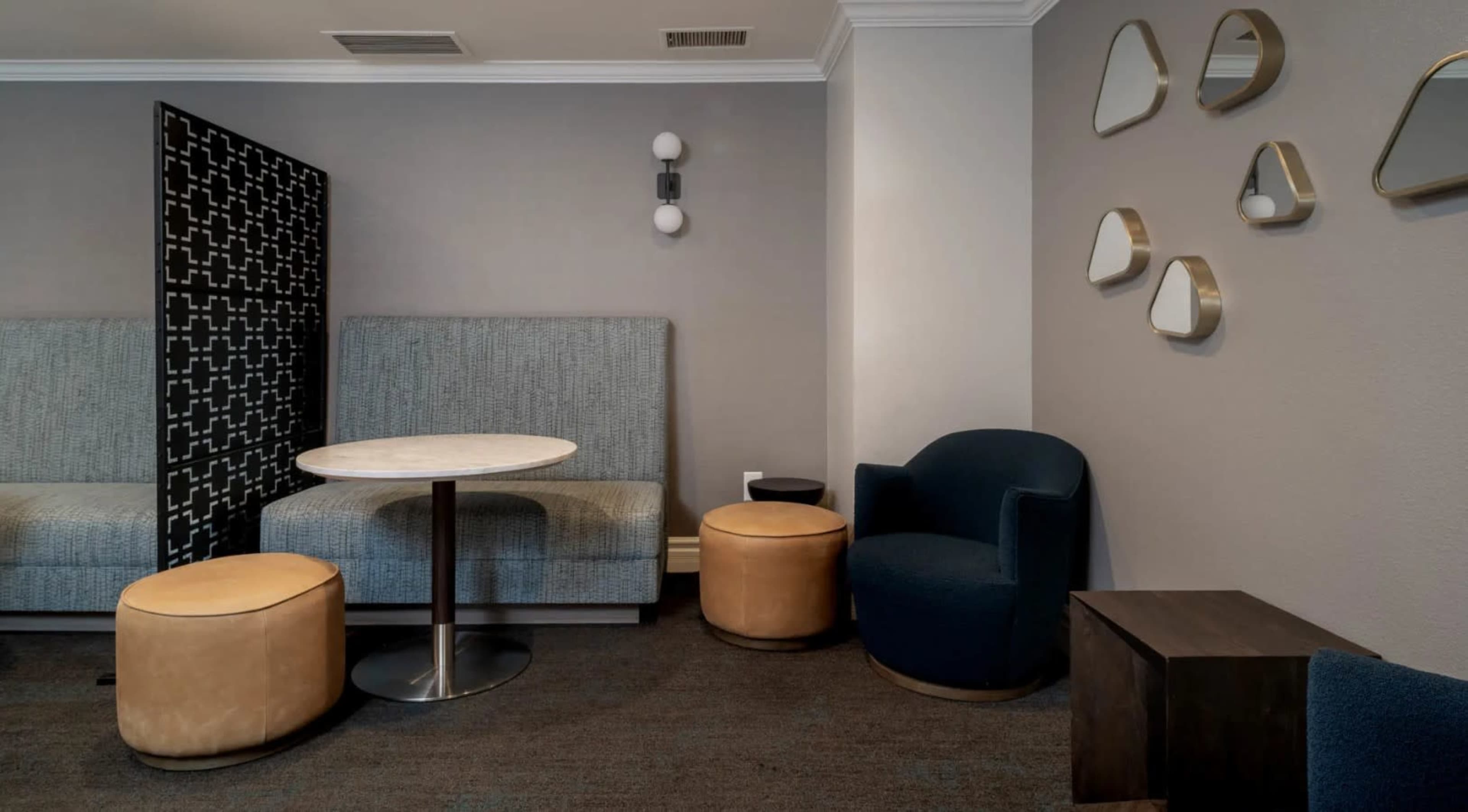 The image shows a modern lounge area featuring a circular table surrounded by upholstered seating and decorative mirrors on the wall.