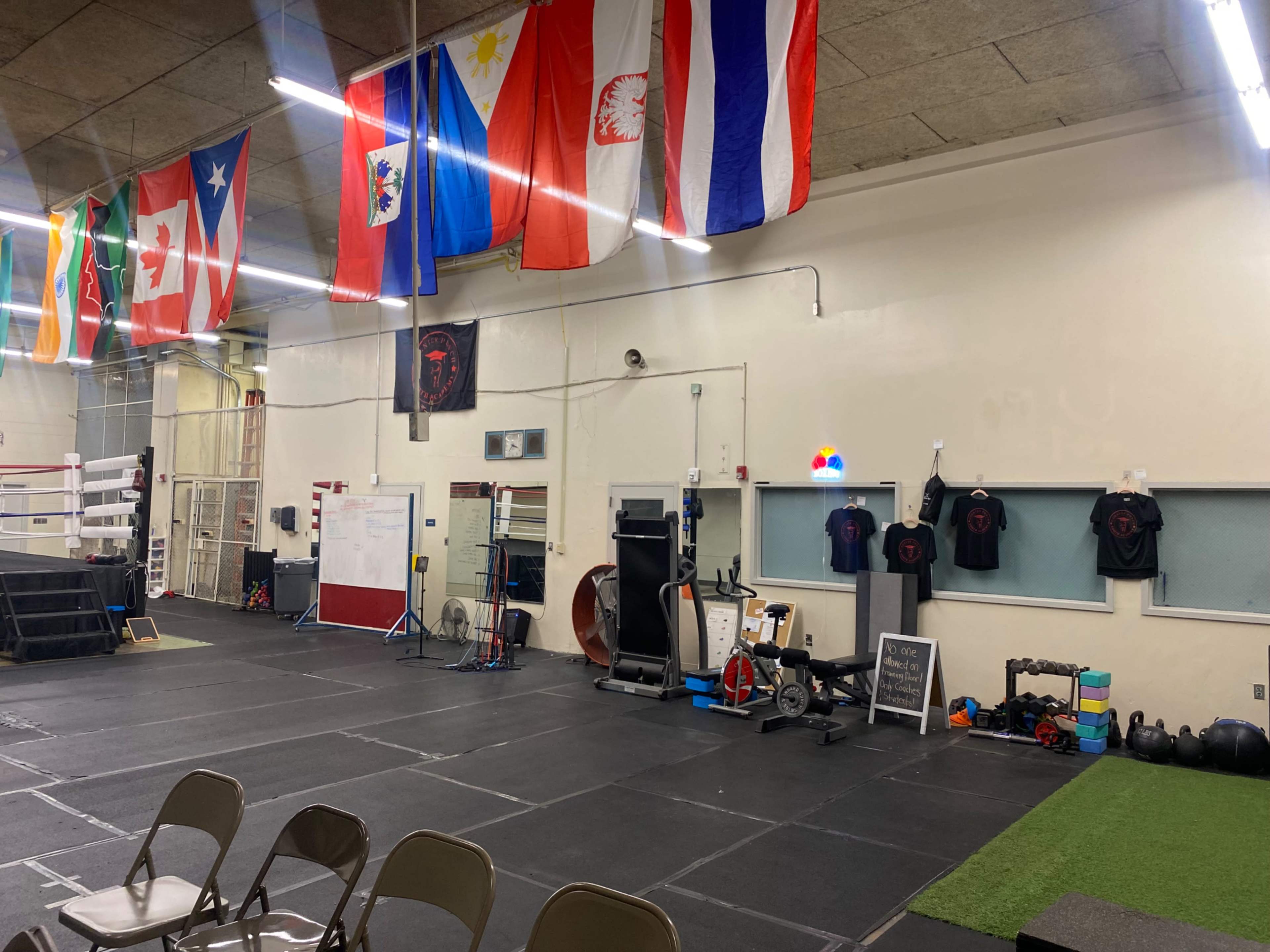The image shows an indoor training facility with various flags hanging from the ceiling, gym equipment, and workout gear displayed on the walls.