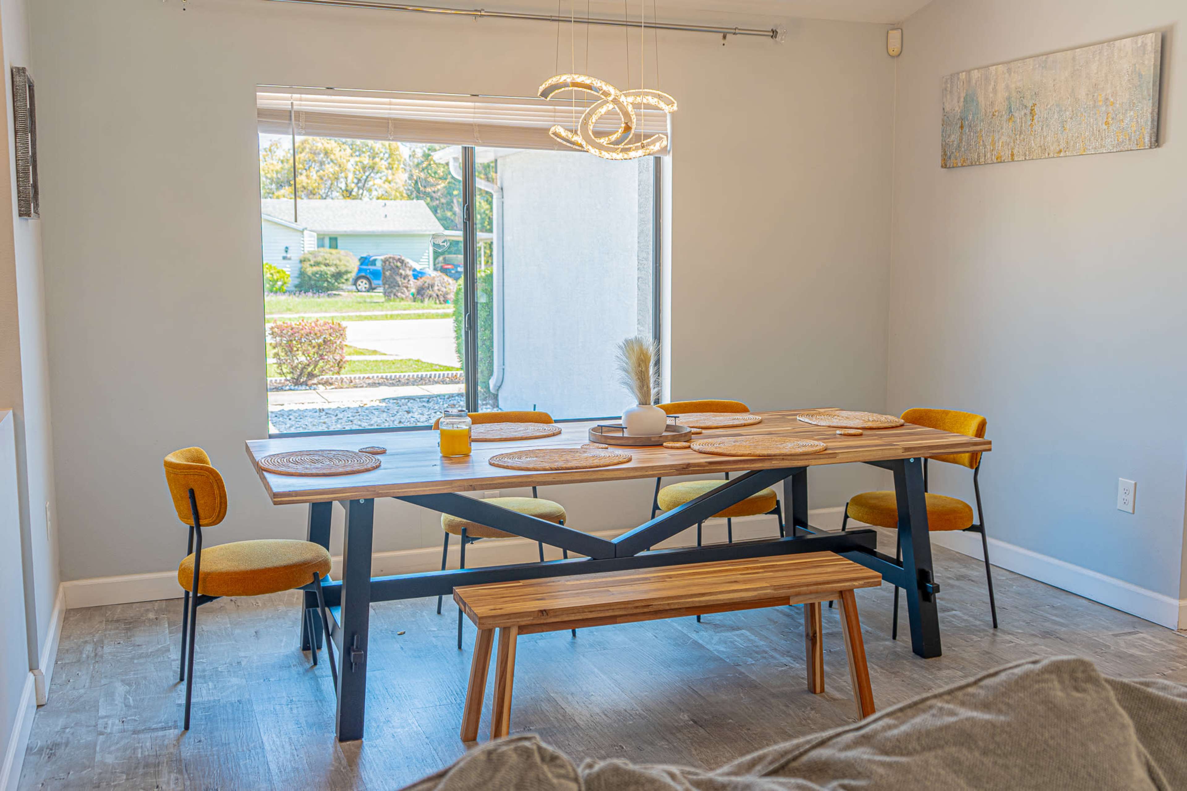 A modern dining area features a wooden table surrounded by yellow chairs and a bench, with a window providing natural light.