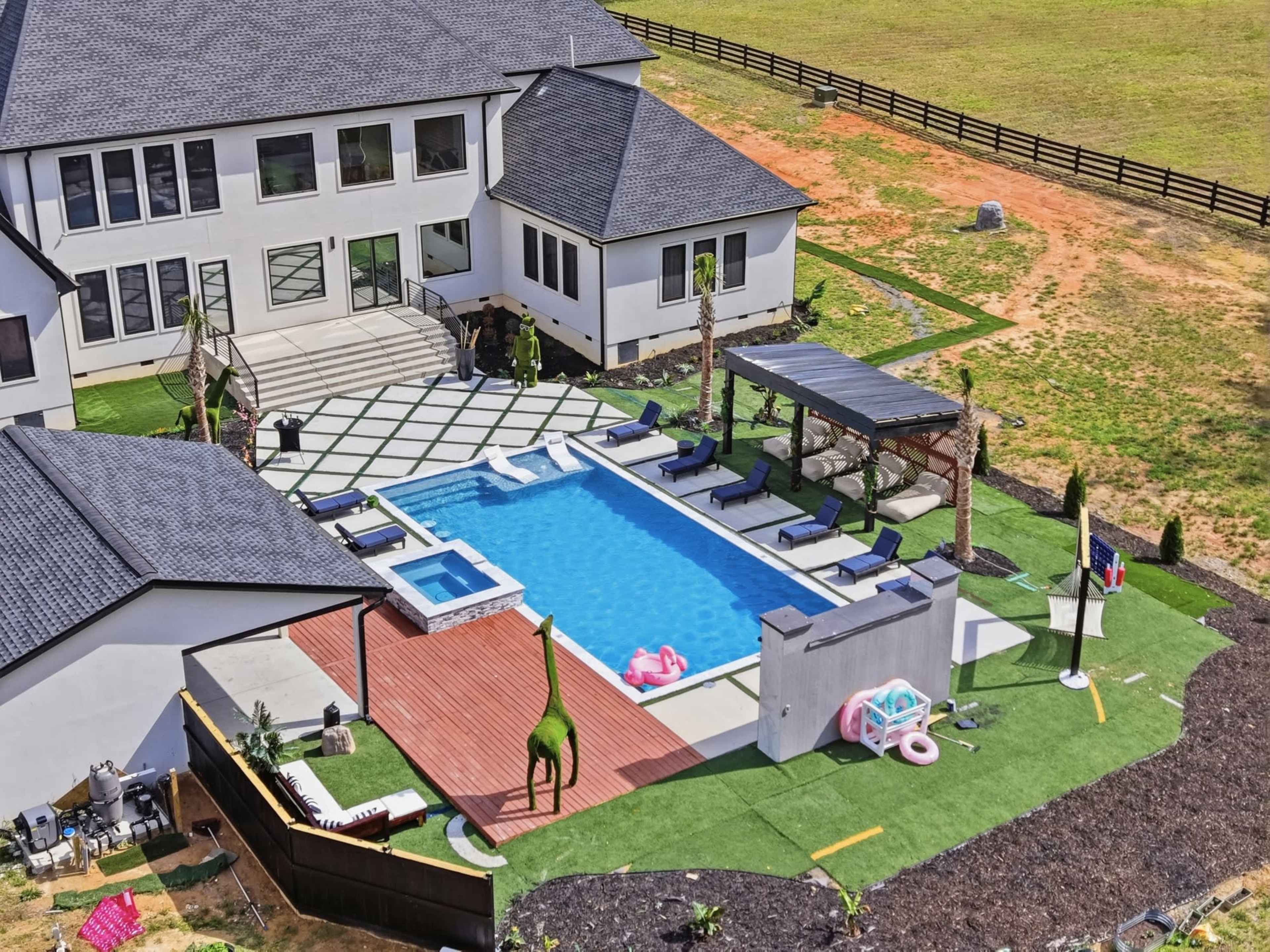 The image shows a large, modern house with an outdoor pool area featuring lounge chairs, a hot tub, and a grassy lawn surrounded by a wooden fence.