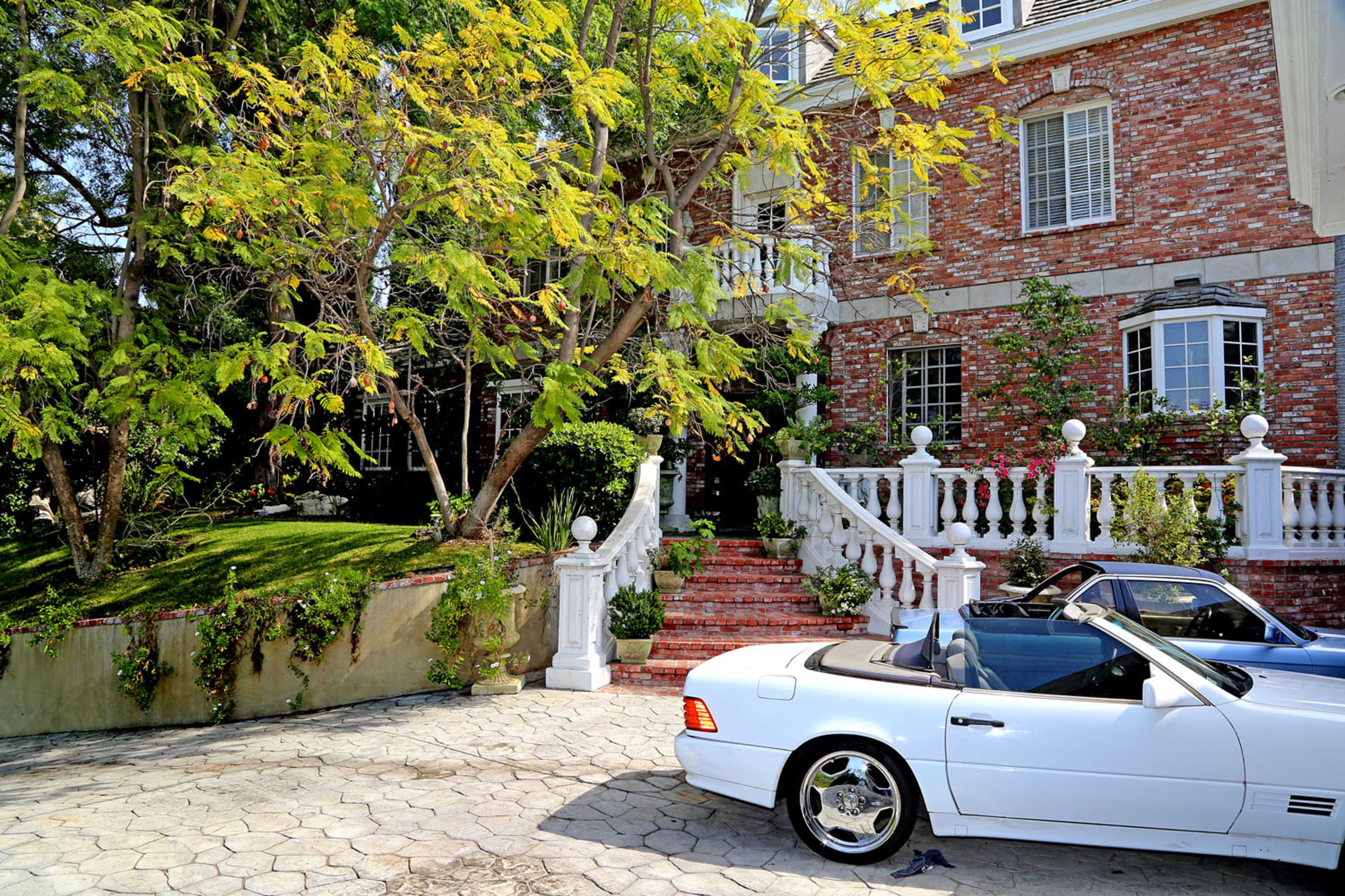 A white convertible car is parked in front of a brick mansion with a landscaped yard and a staircase flanked by white columns.