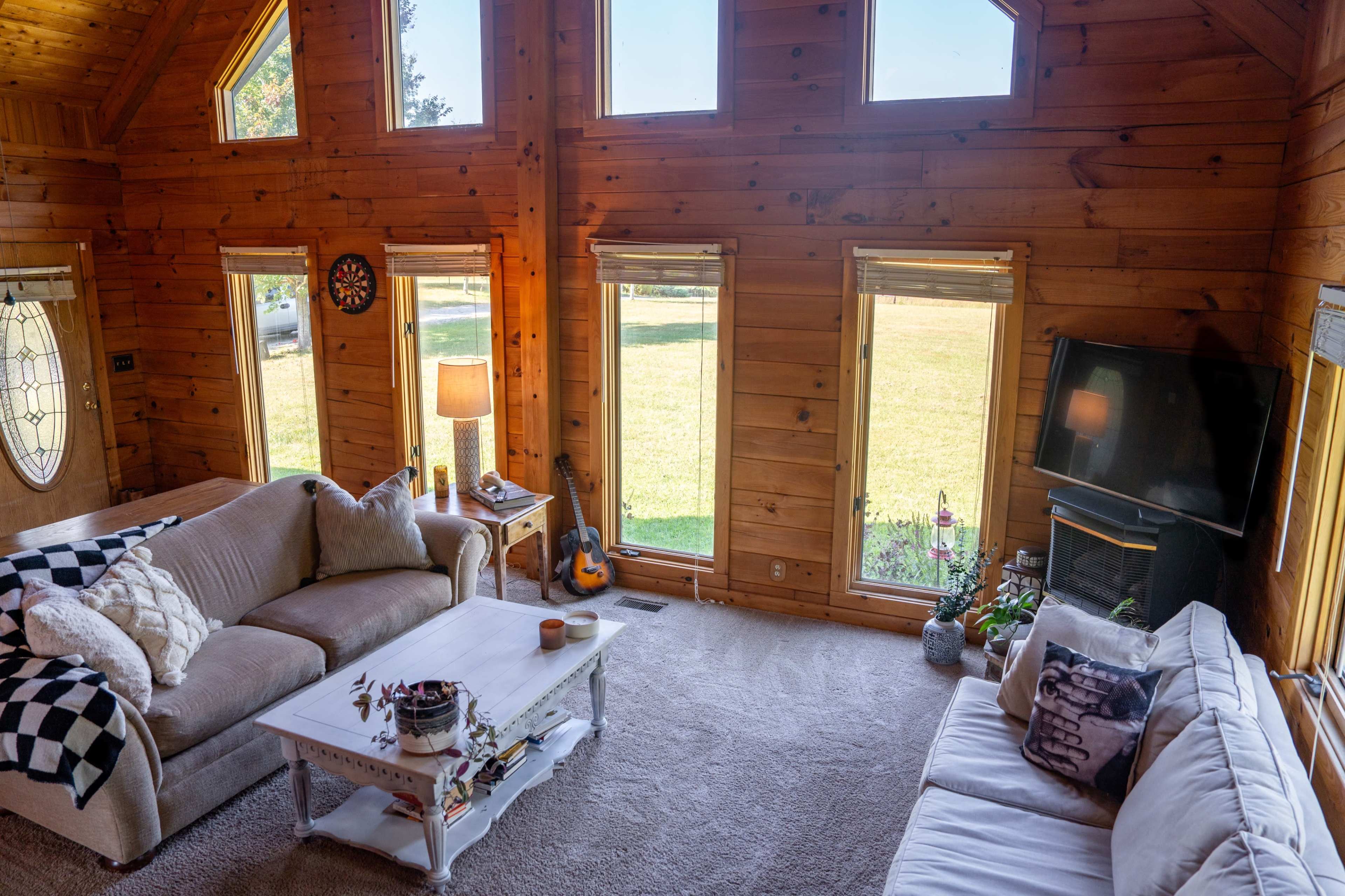 A wooden cabin living room with large windows, a coffee table, two sofas, and a guitar resting in the corner.