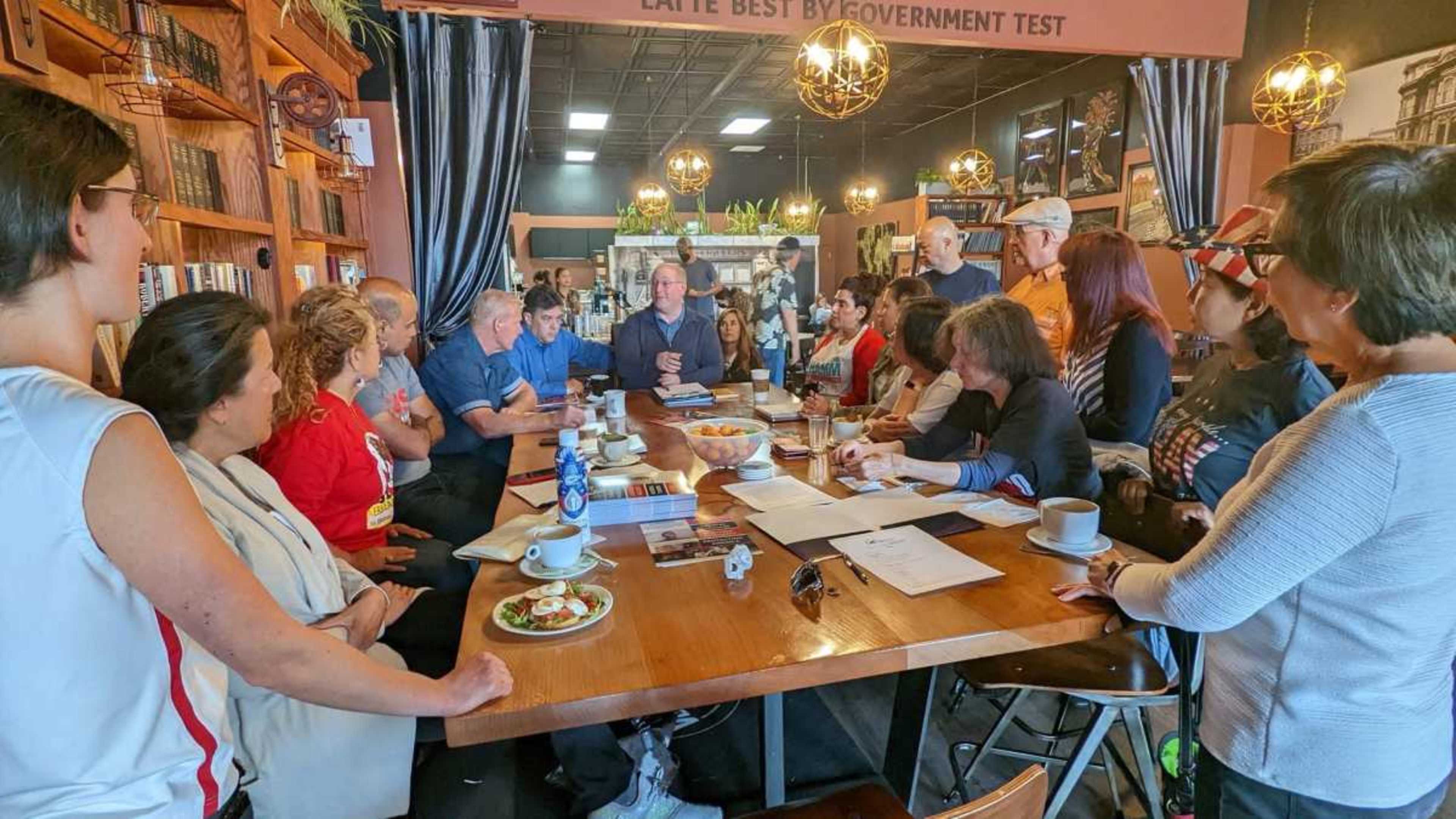 A group of people sits around a large table in a cafe, engaged in conversation and discussion.