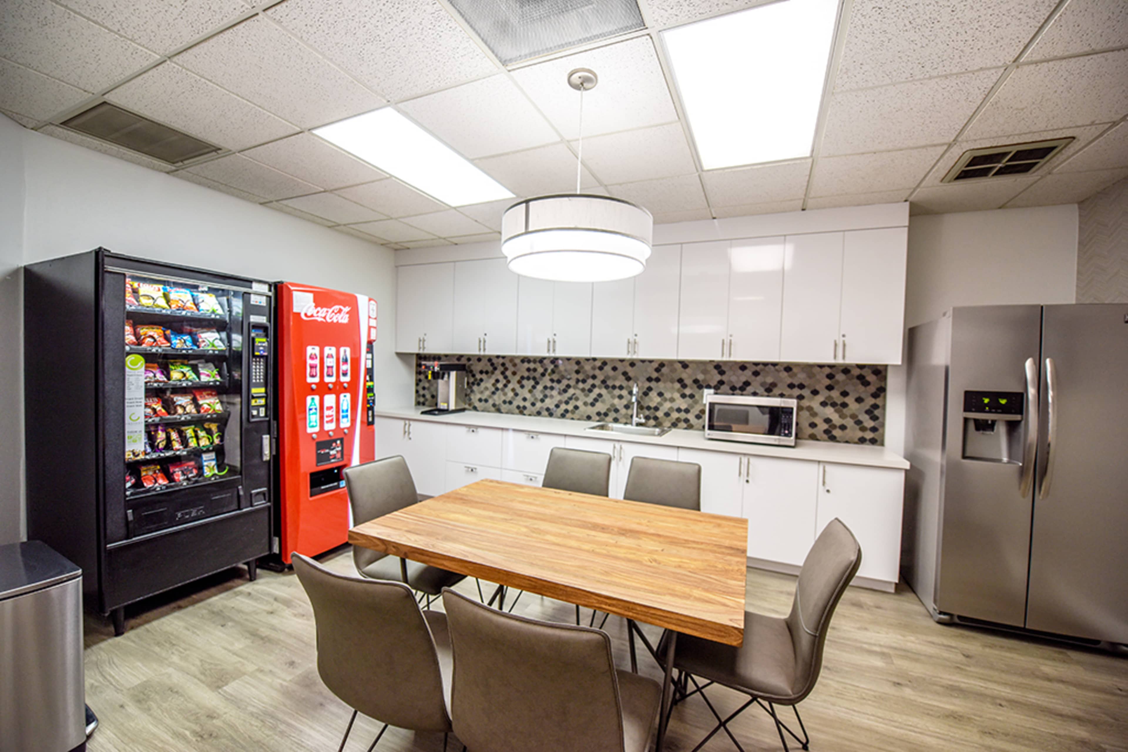A modern kitchen area with a wooden dining table surrounded by six chairs, a large vending machine, a Coca-Cola vending machine, a microwave, and a stainless steel refrigerator.