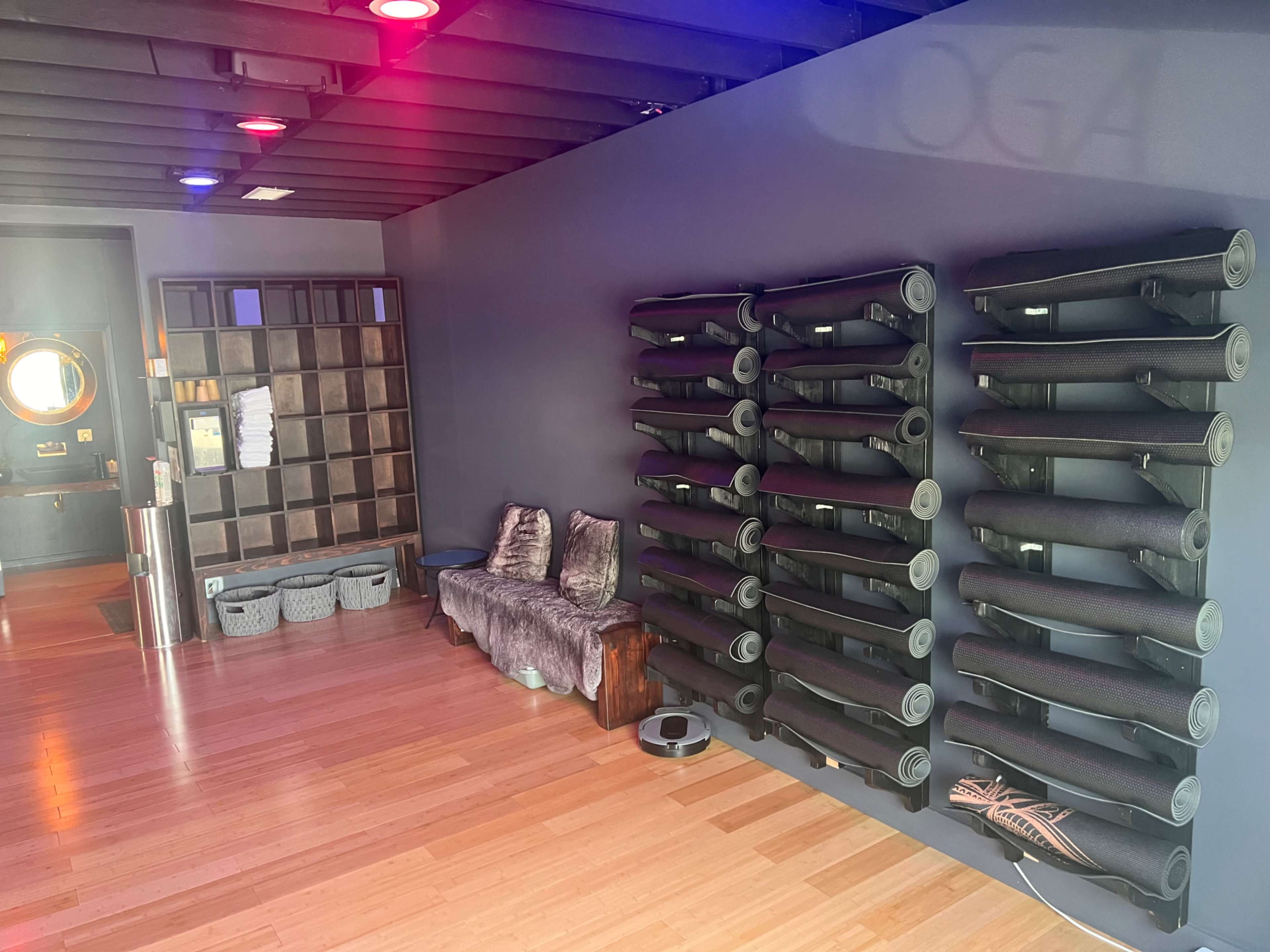 The image shows a yoga studio interior with neatly stacked yoga mats on a wall, a seating area with cushions, and cubbies for storage.