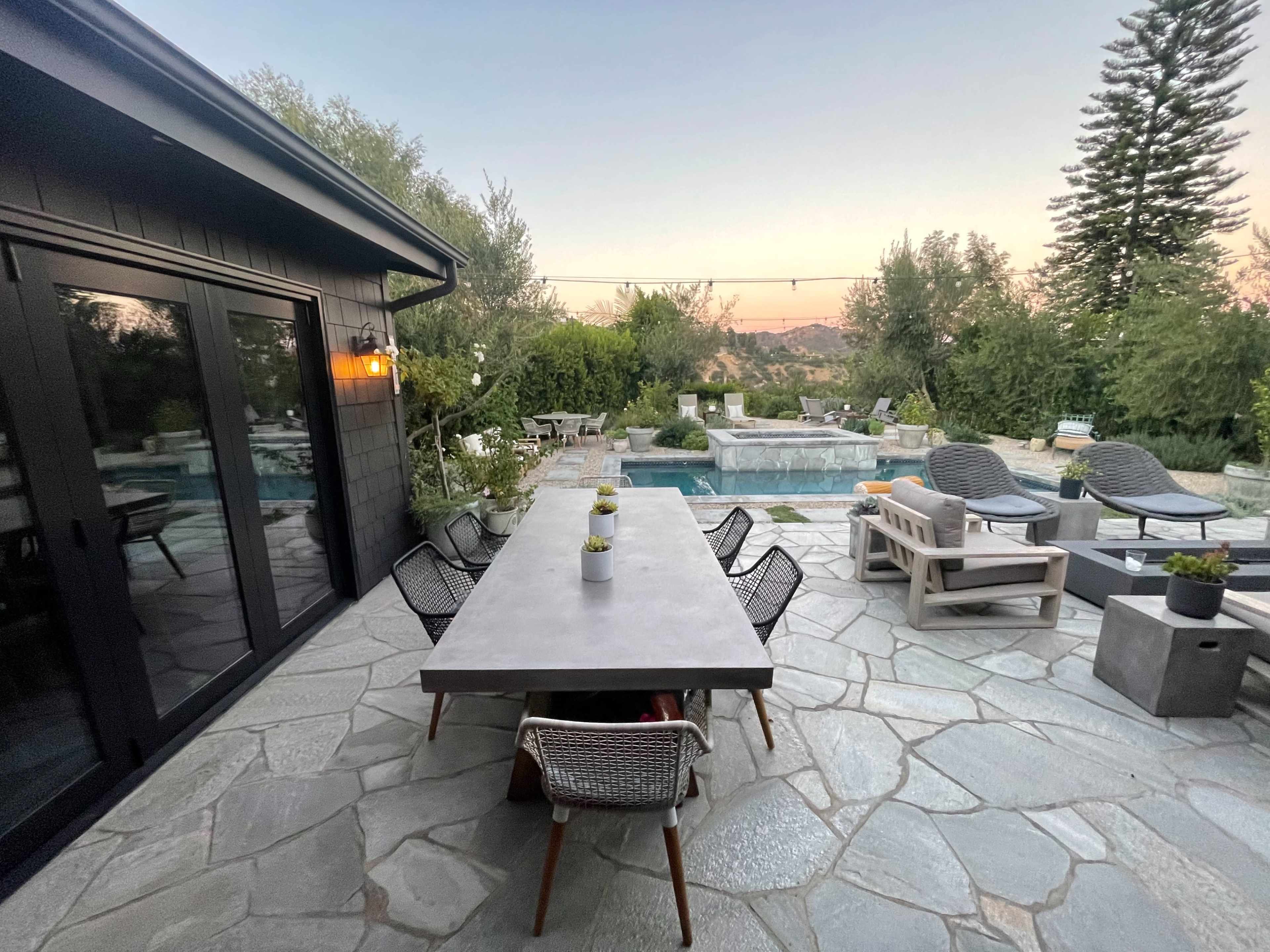 A modern outdoor patio features a large stone table surrounded by chairs, with a pool and landscaped garden in the background.