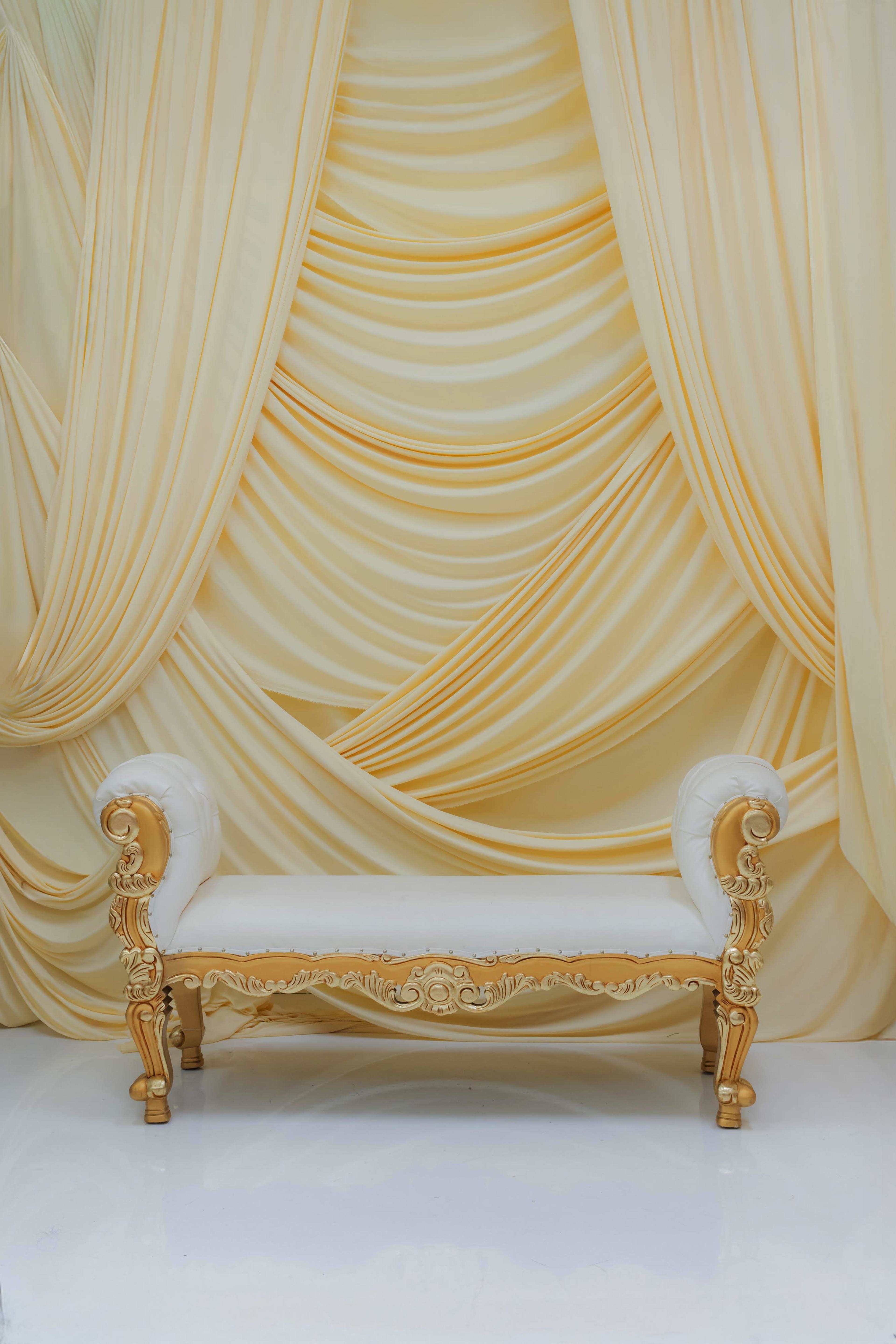 The image features an elegant white bench with ornate golden details set against a backdrop of draped yellow fabric.
