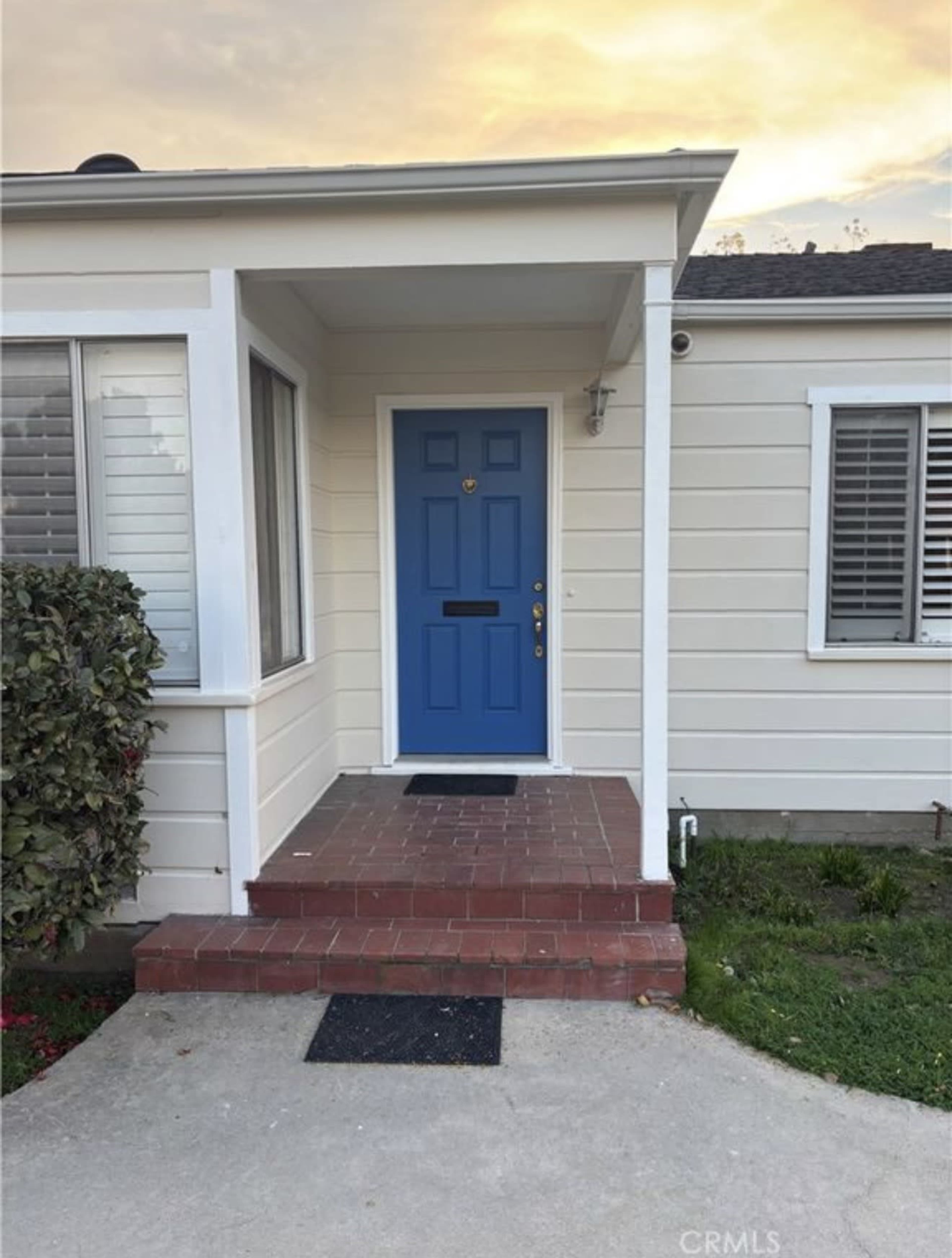 The image shows a house entrance with a bright blue door, brick steps, and a small garden area adjacent to the pathway.