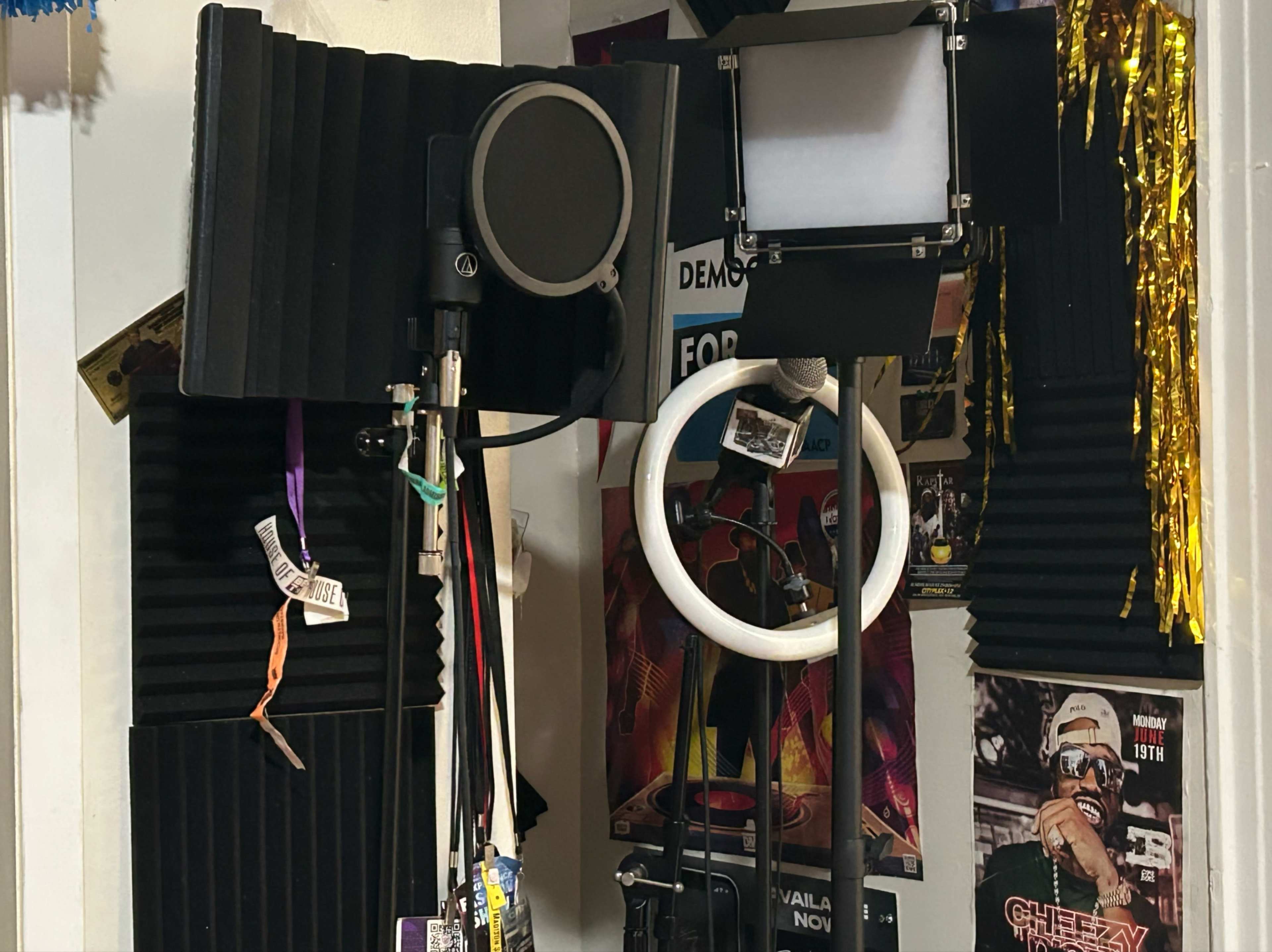 The photo shows a small recording space equipped with soundproofing panels, a microphone, and lighting equipment surrounded by various posters.