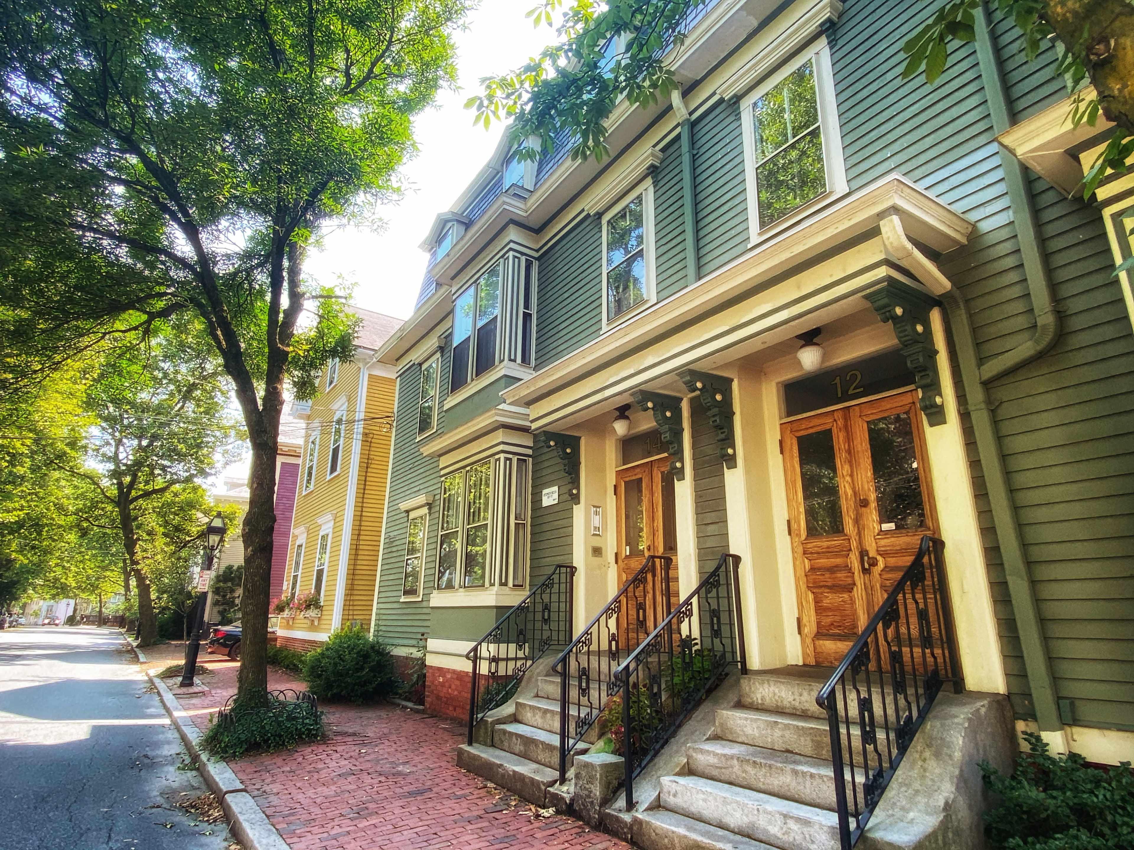 A row of green townhouses with wooden front doors and stairs, set on a tree-lined street.